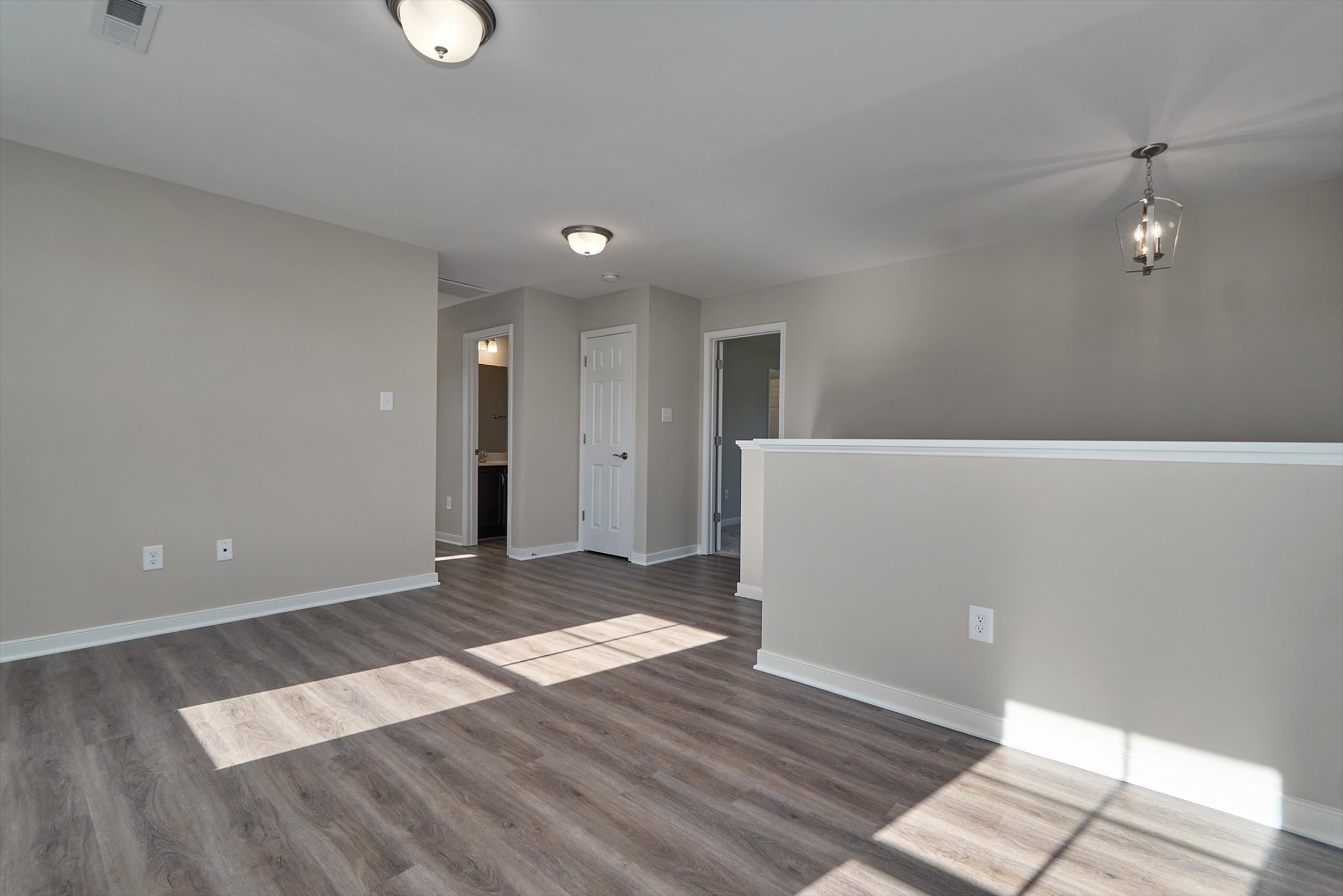 A well-lit empty room with light gray walls, dark wood flooring, and modern light fixtures. The space features a doorway, a half-wall, and several power outlets. Natural light enters from the left.