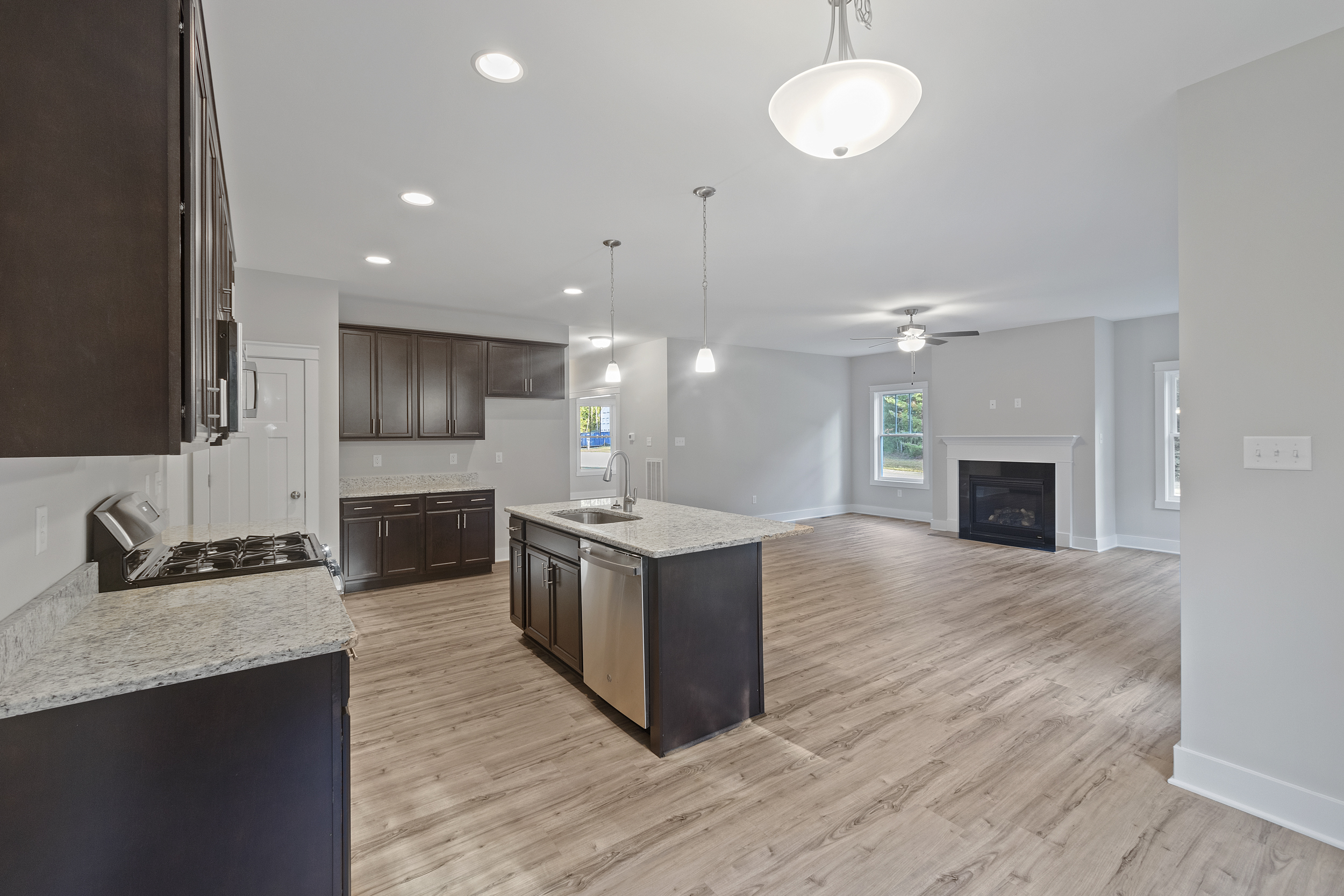 A modern, open-concept kitchen and living area with hardwood floors, dark wood cabinetry, a center island with a stainless steel sink, pendant lighting, and a fireplace.