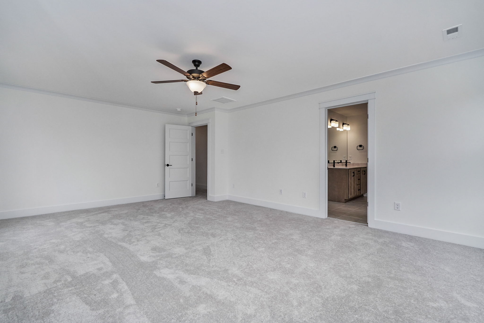 A spacious, carpeted room with a ceiling fan, white walls, a door leading to an adjoining room, and another door leading to a bathroom with a double sink vanity.