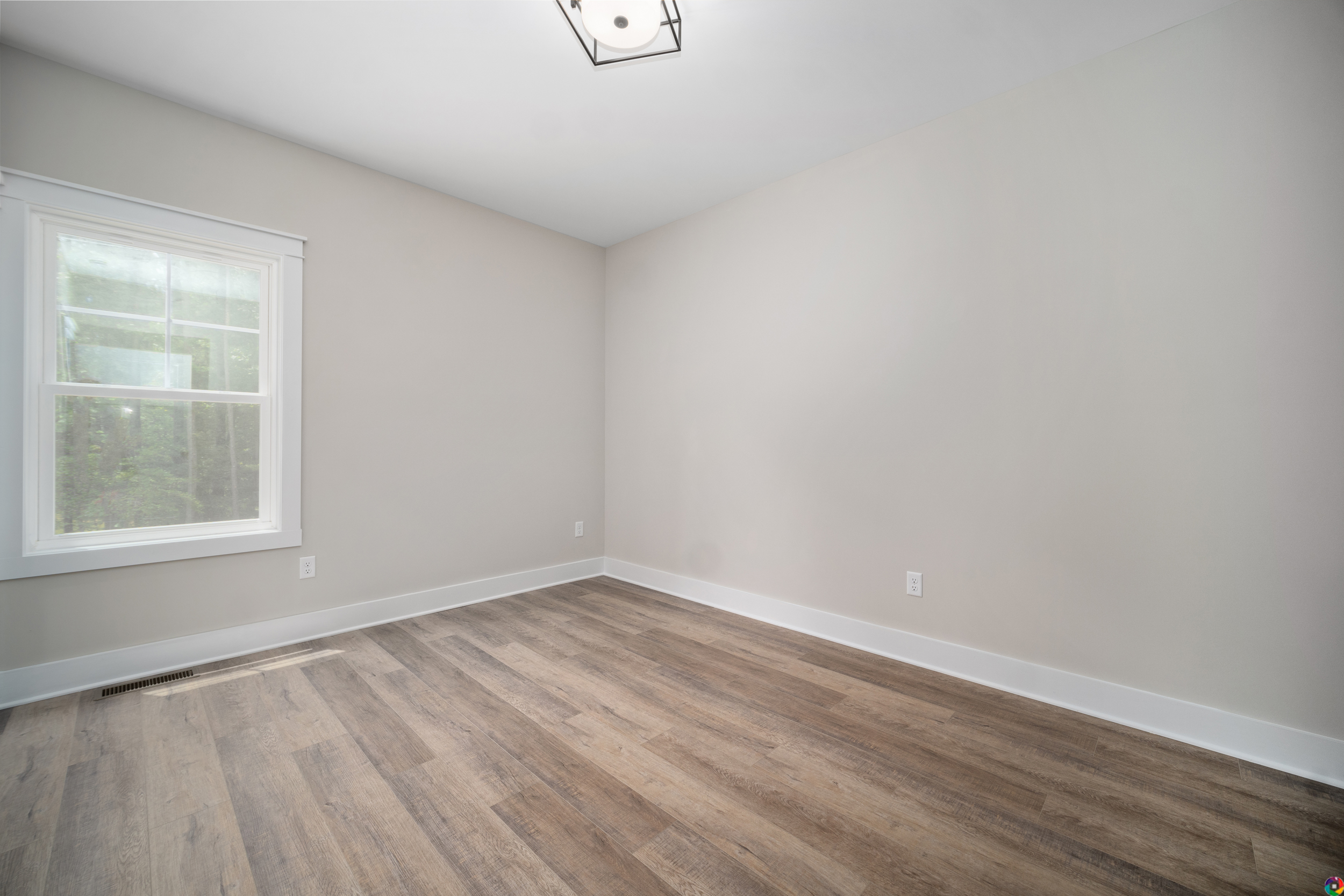 A minimalistic, empty room with light gray walls, a single window with white trim, and hardwood flooring, illuminated by a ceiling light.