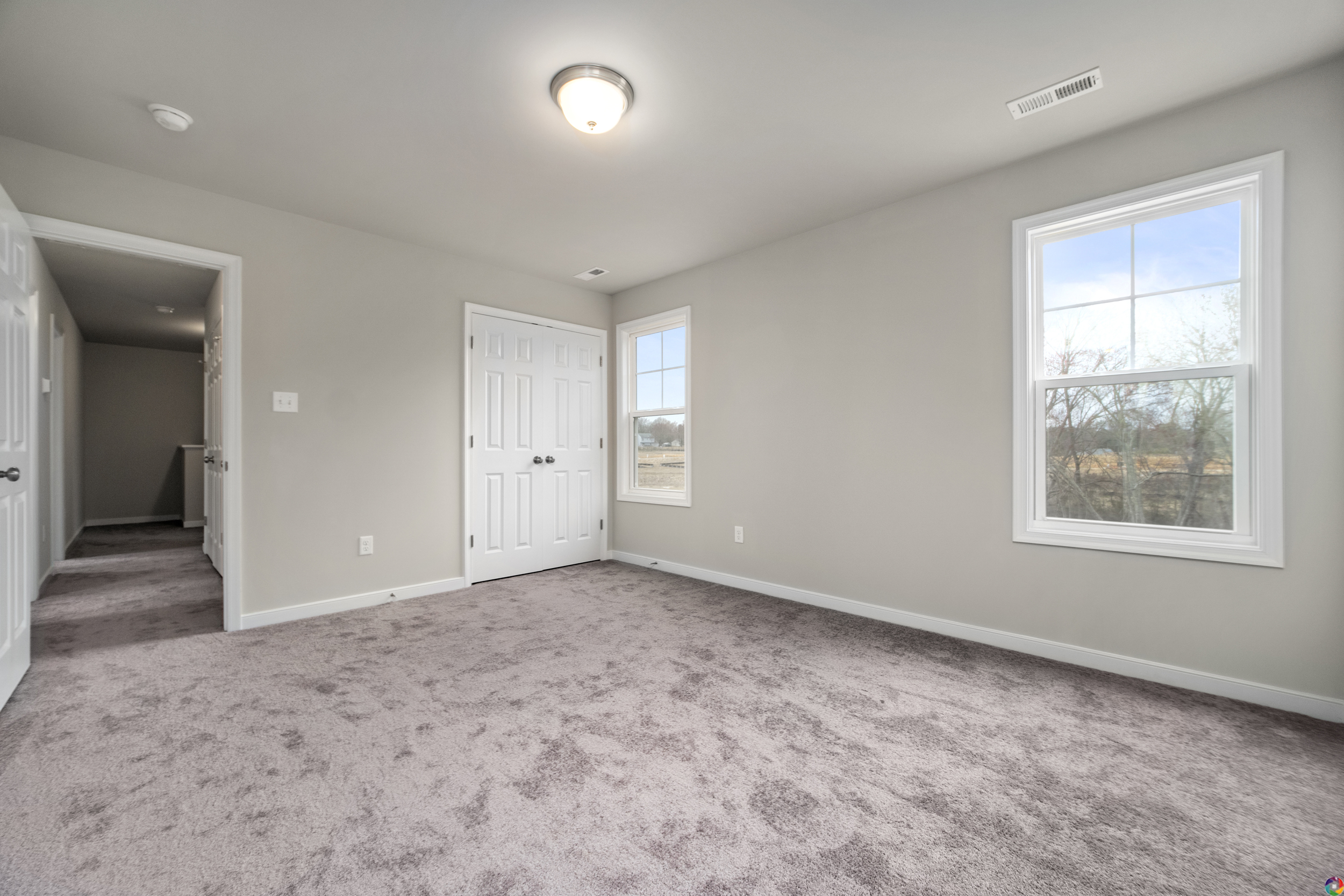 A vacant, carpeted room with beige walls, a ceiling light, two windows, a closed closet with double doors, and an open door leading to a hallway.