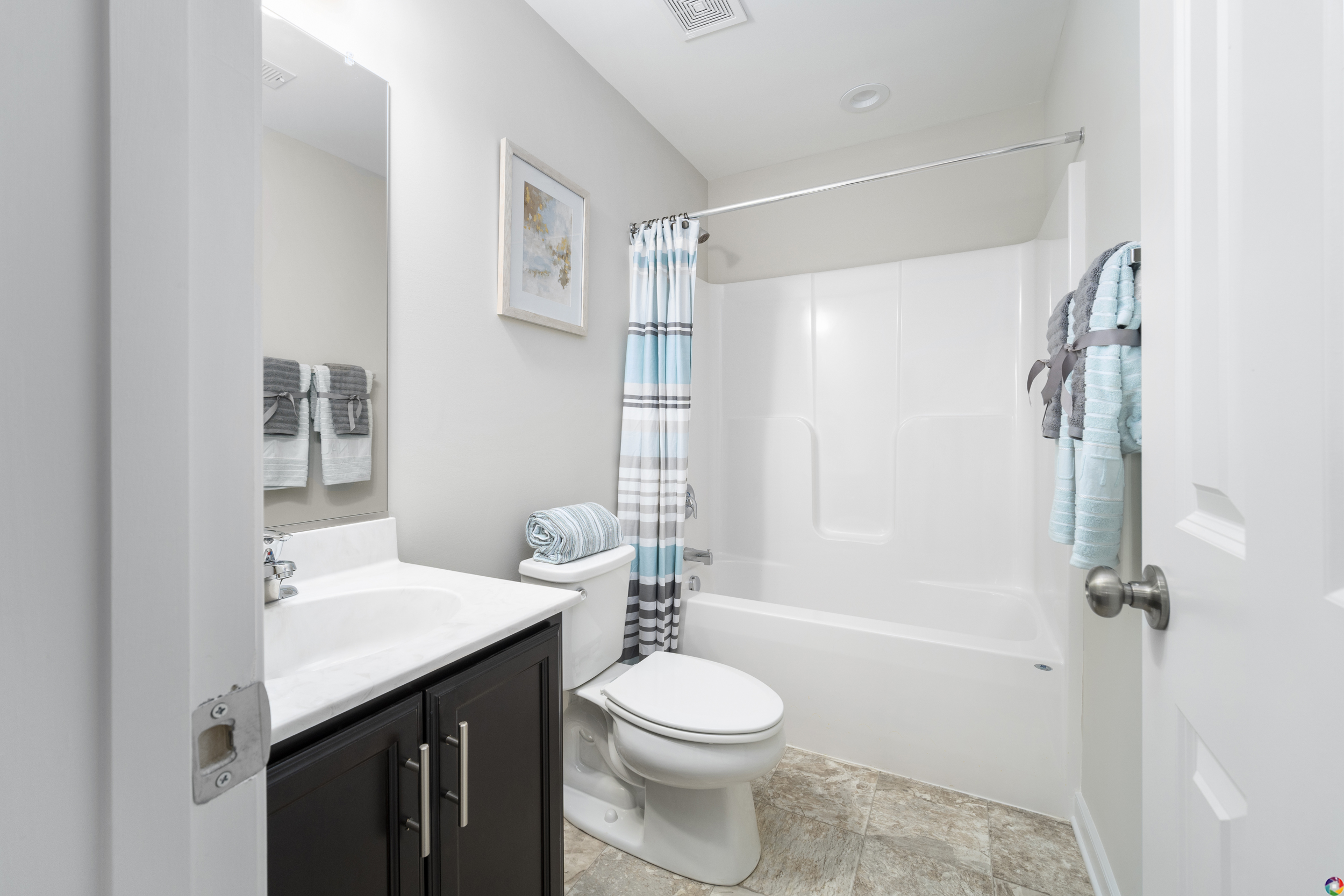 A modern, clean bathroom featuring a white tub with a shower, a toilet, a vanity with a sink, and neatly hung towels. decor includes framed beach-themed art on the walls.