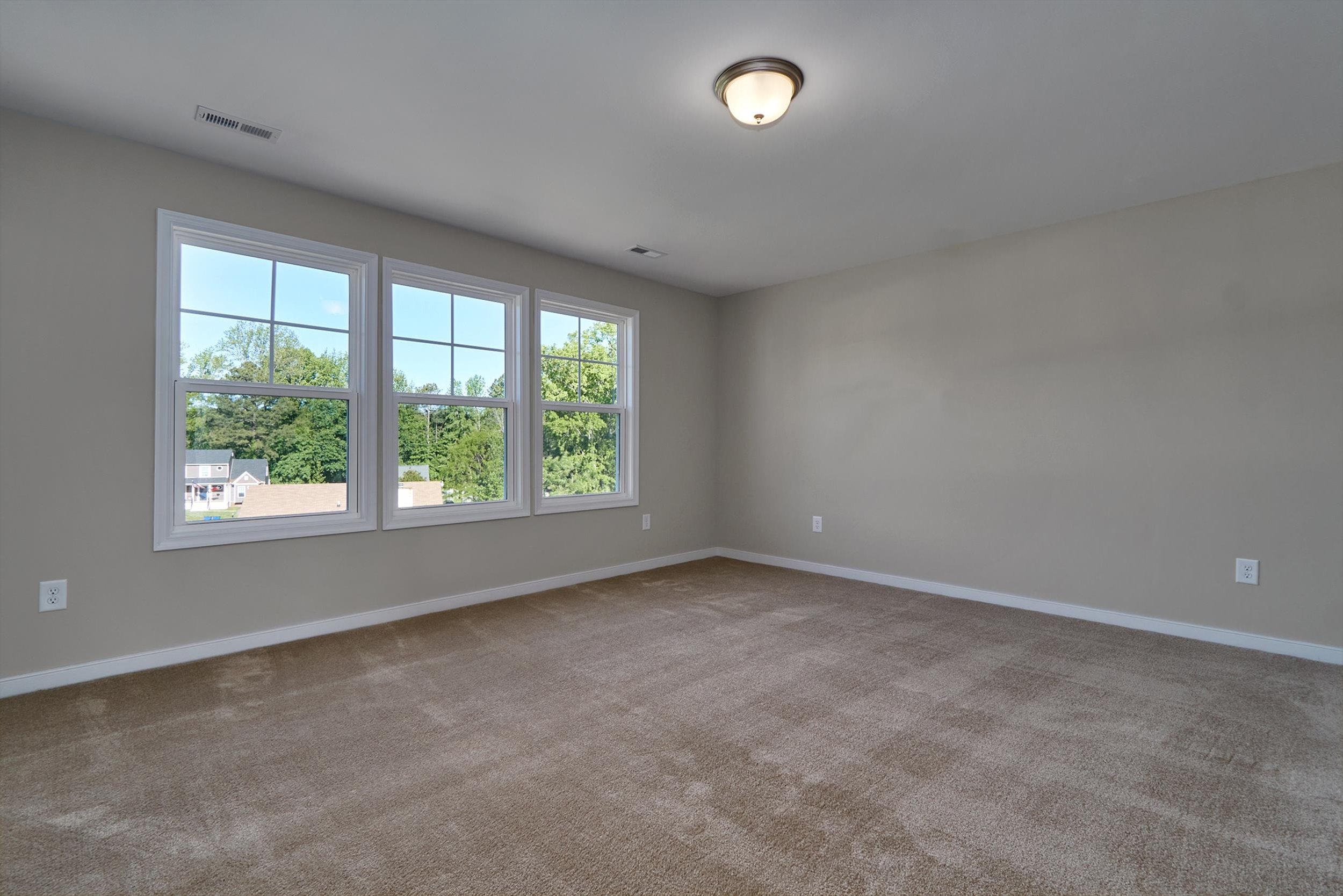 A spacious, empty room with beige carpet, light gray walls, and a ceiling light fixture. Three large windows allow natural light to enter, showing a view of greenery outside.