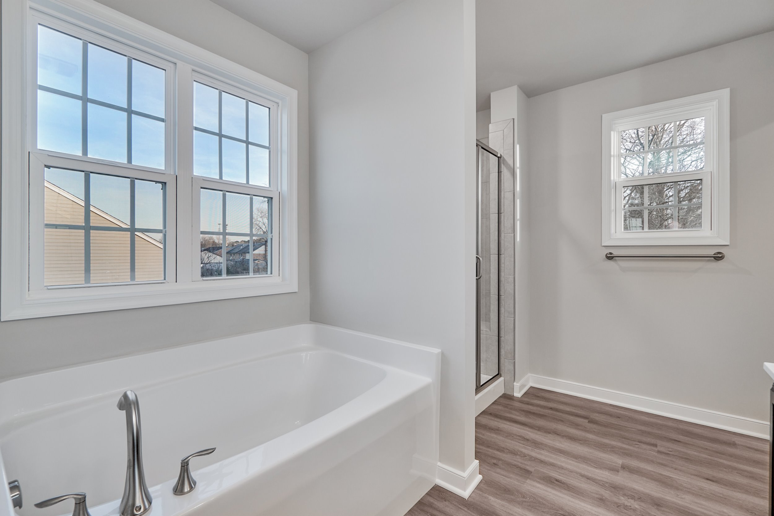 A modern bathroom featuring a large bathtub, glass-enclosed shower, and two windows. The floor is made of light-colored wood.