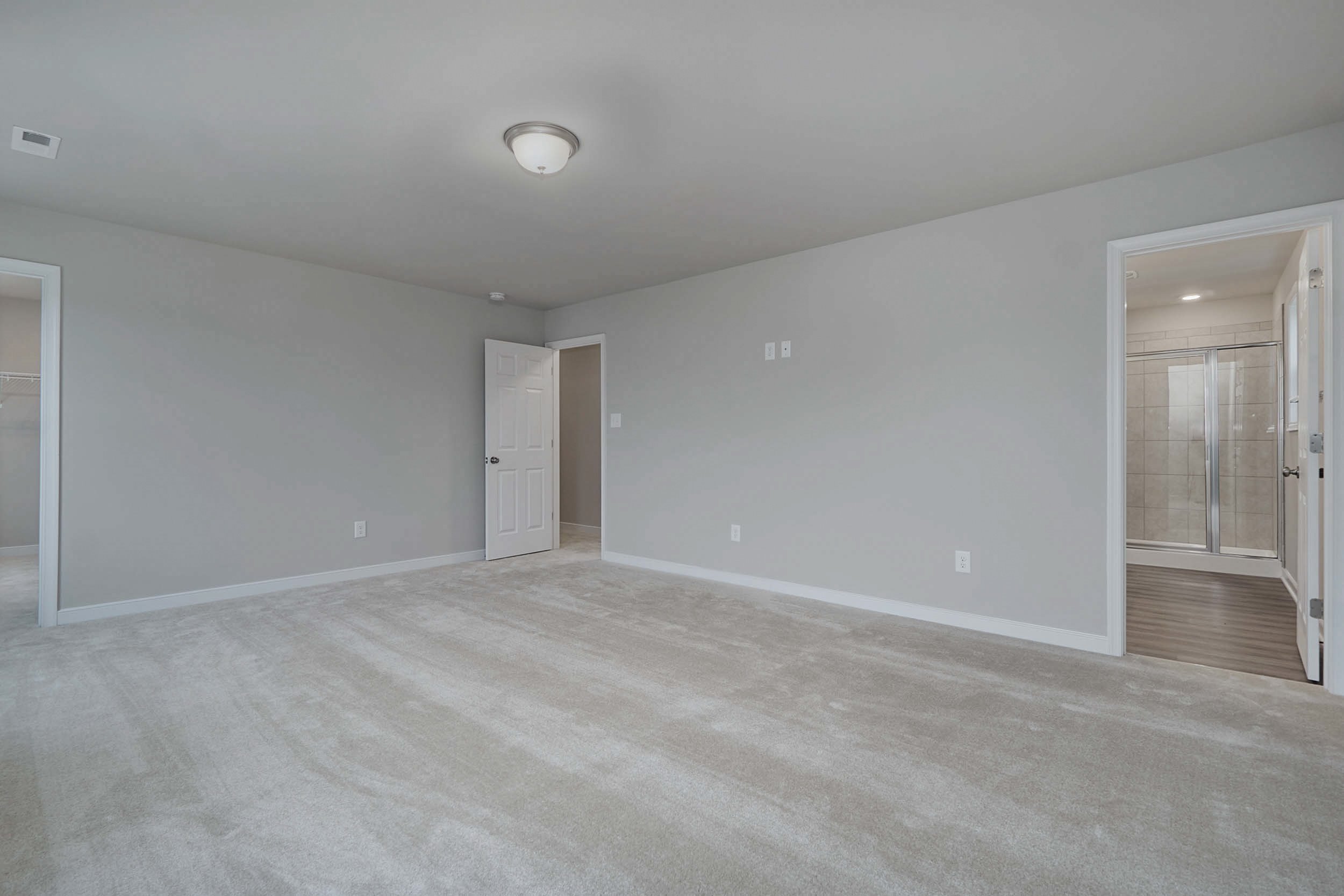 A spacious, empty room with light gray walls and carpet, two open doorways, one leading to a closet and the other to a bathroom with a glass shower. Ceiling light fixture in center.