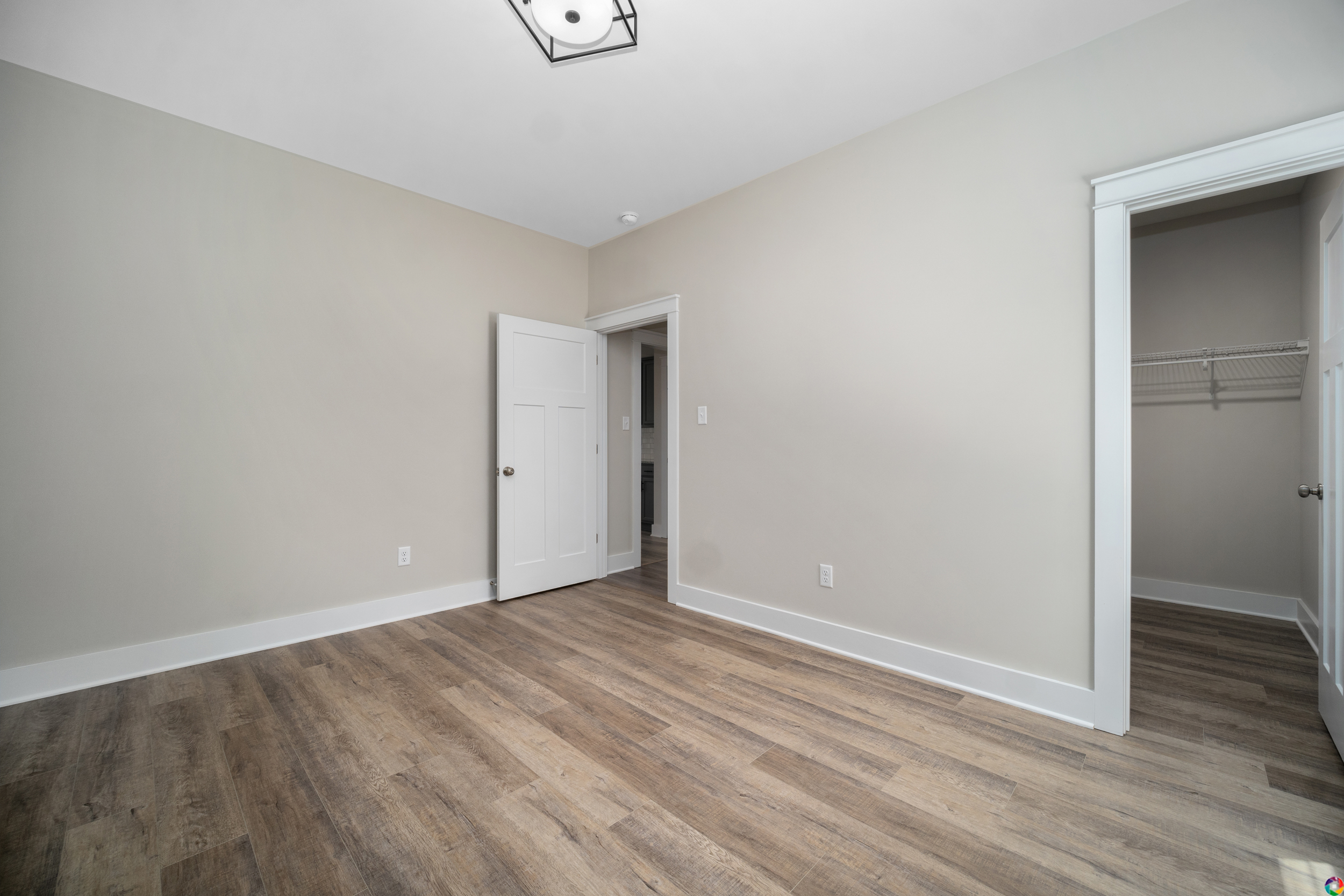 An empty room with light gray walls, a white door, a walk-in closet, and light wood flooring. There is a light fixture on the ceiling.