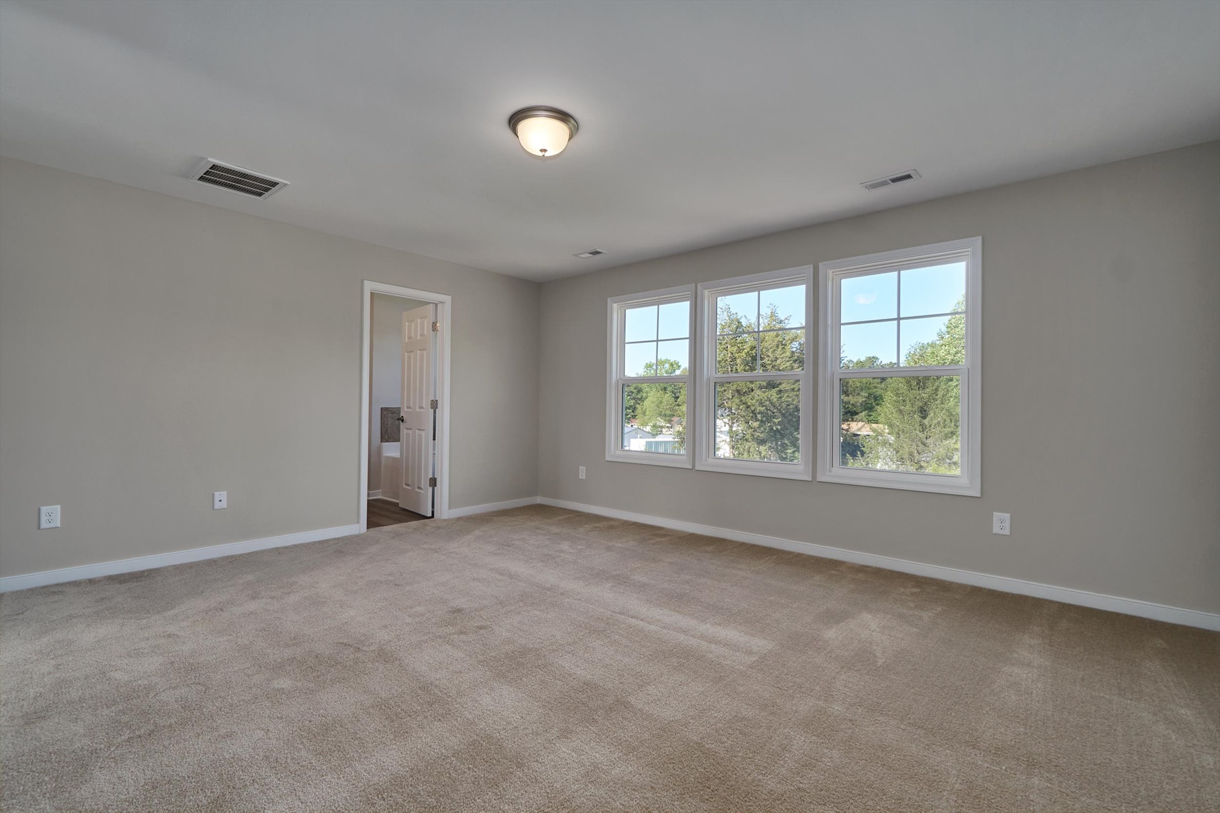 A spacious, empty room with beige carpet, light gray walls, three windows, a ceiling light fixture, and a doorway leading to another room.