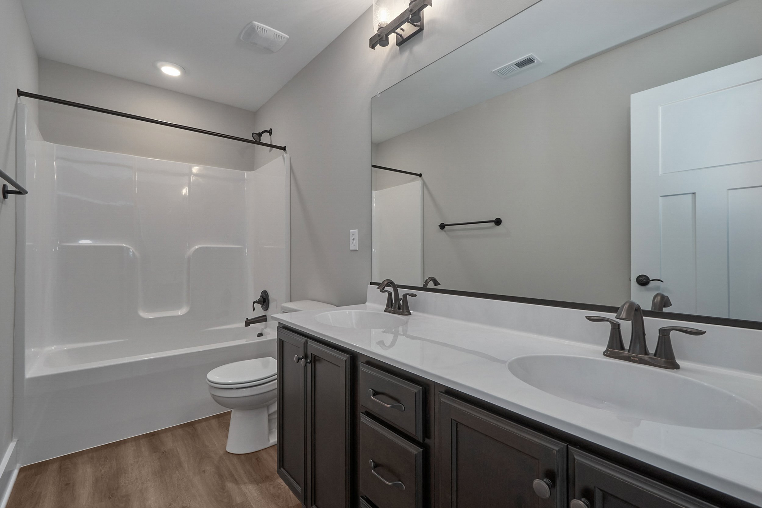 Modern bathroom featuring a double sink vanity with dark cabinets, large mirror, bathtub with shower, white walls, and wood-look flooring.