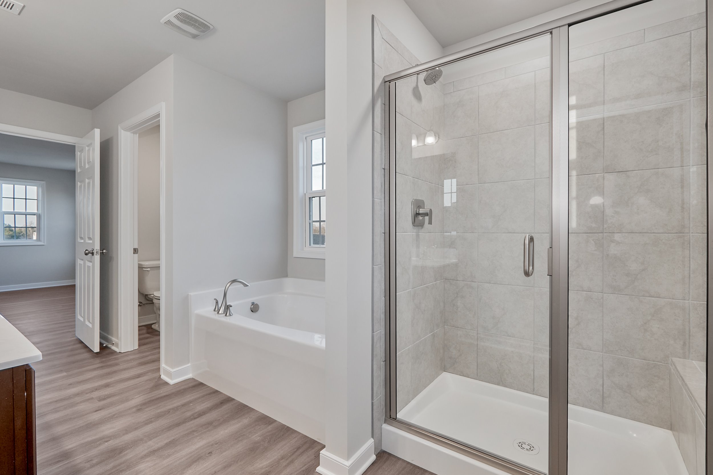 Modern bathroom featuring a glass-enclosed shower, a large bathtub, and a doorway leading to a toilet area. The room has light gray walls and wood flooring.