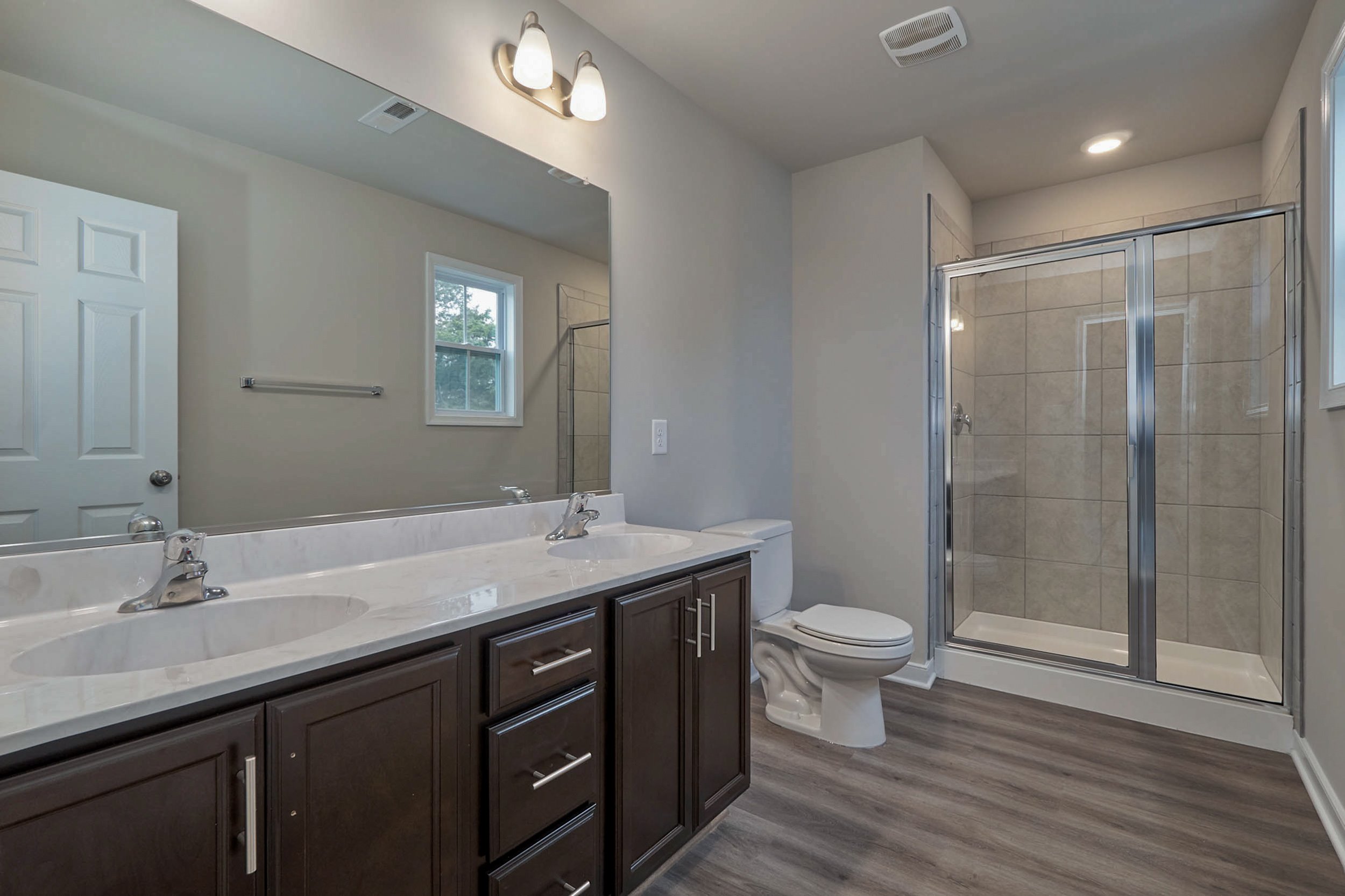 A modern bathroom featuring a double sink vanity with dark cabinets, a toilet, a glass-enclosed shower, and wooden flooring.