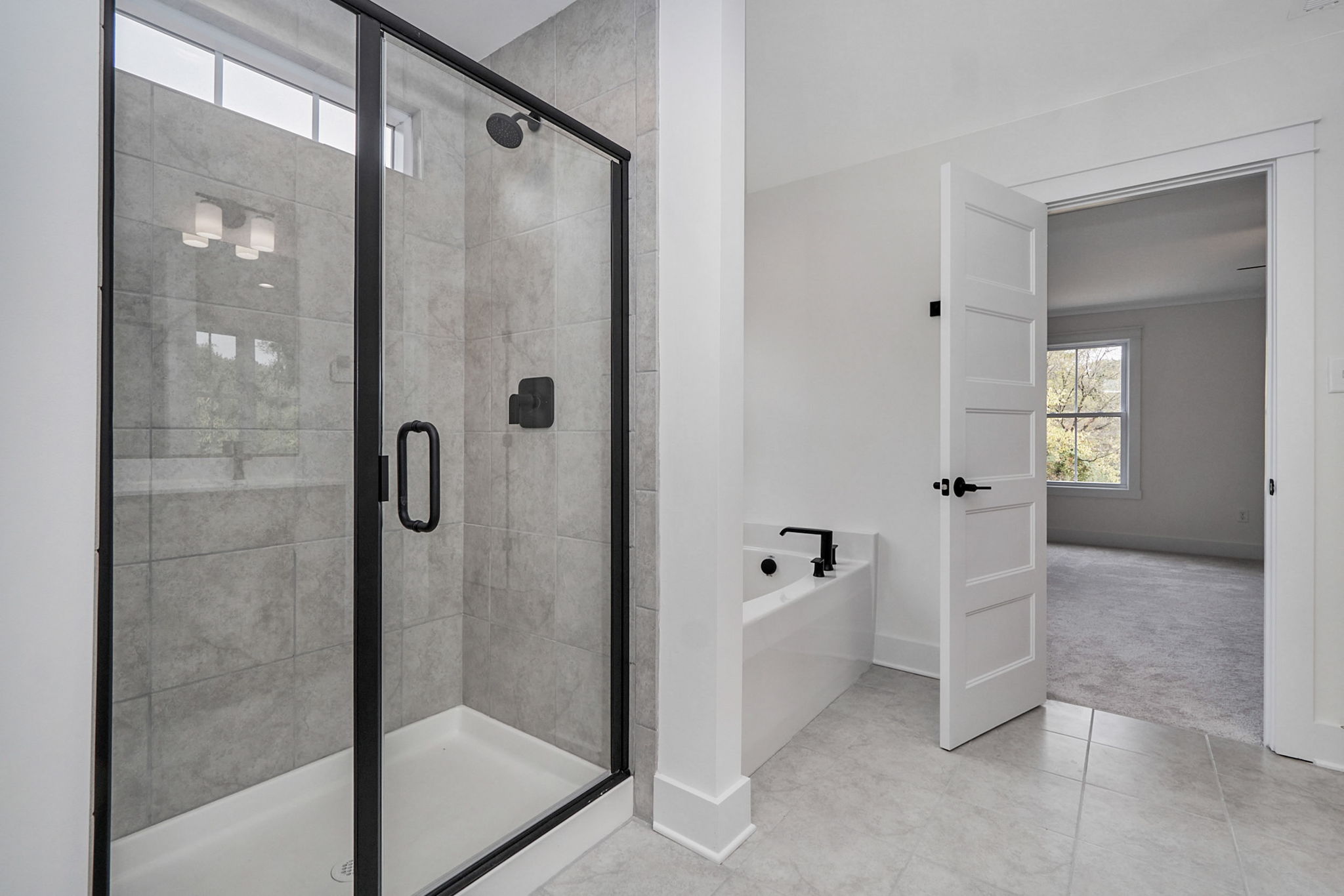 Modern bathroom with a glass-enclosed shower, a bathtub with black fixtures, and an open door leading to a carpeted room with a window.