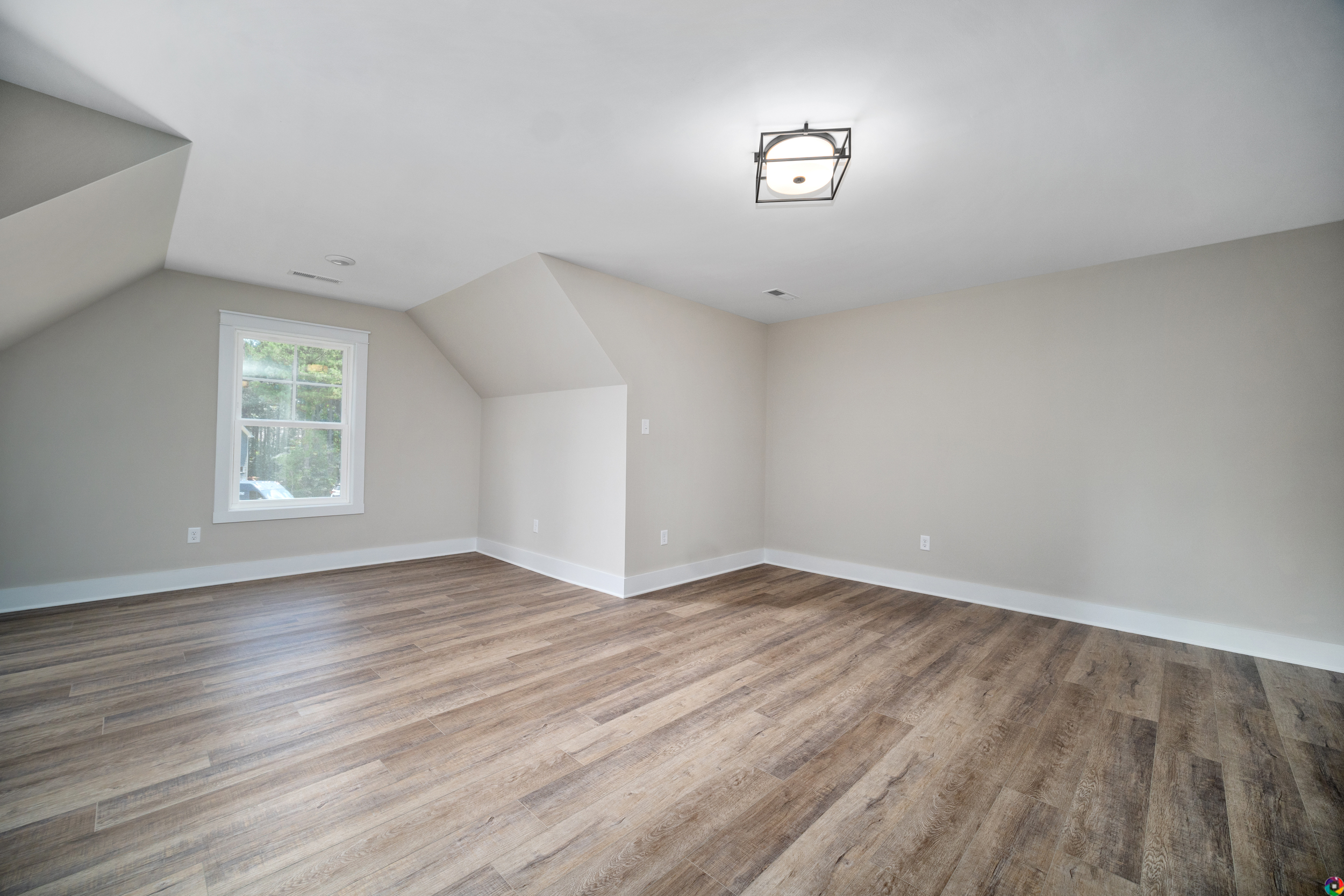 A spacious, empty room with light-colored walls, a window, and wood flooring. It has a modern ceiling light fixture, and angled ceilings on the left side.