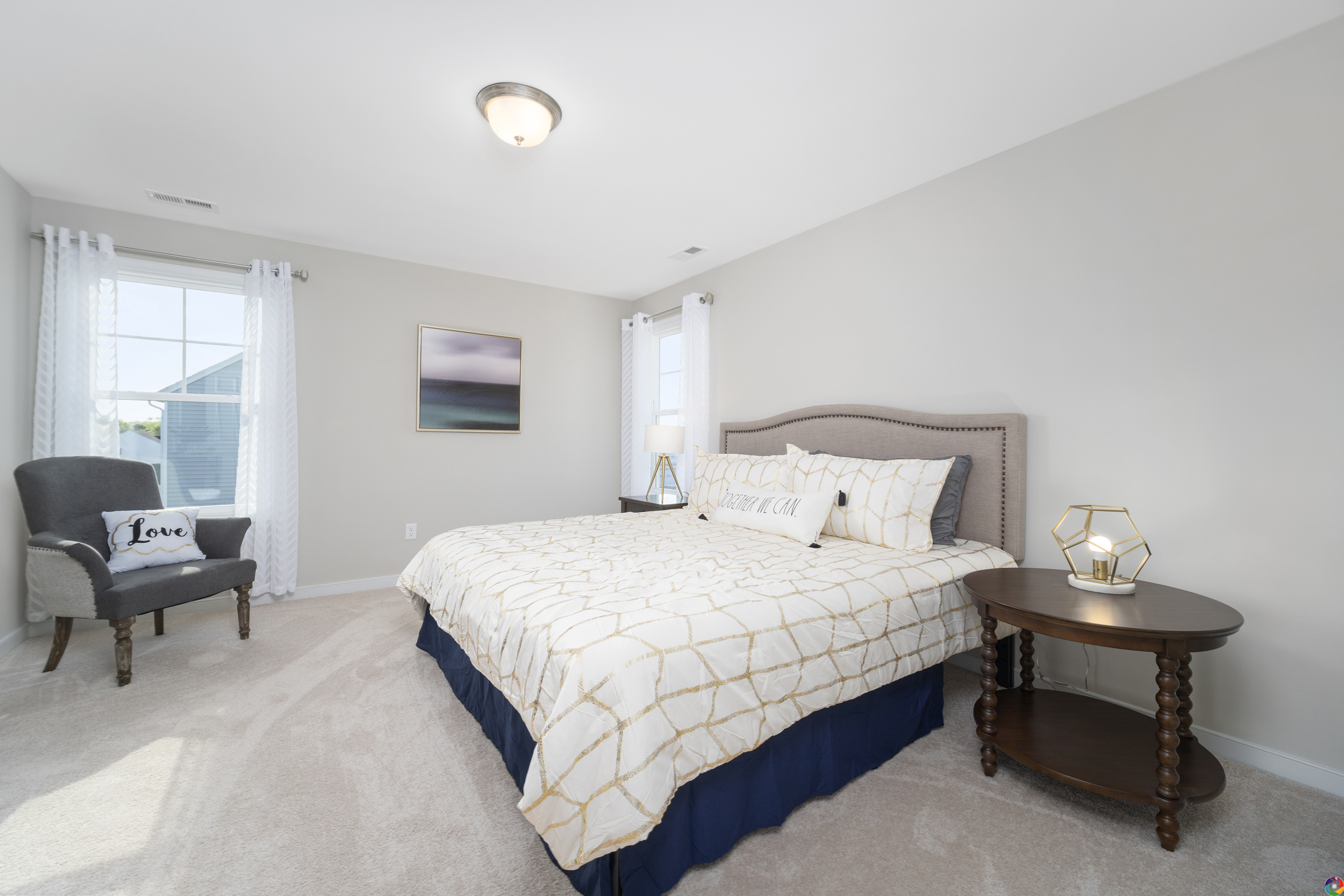 A bedroom with a large bed, patterned bedding, and decorative pillows. A round wooden side table is next to the bed. A gray upholstered chair is in the corner, with sheer curtains on the windows.