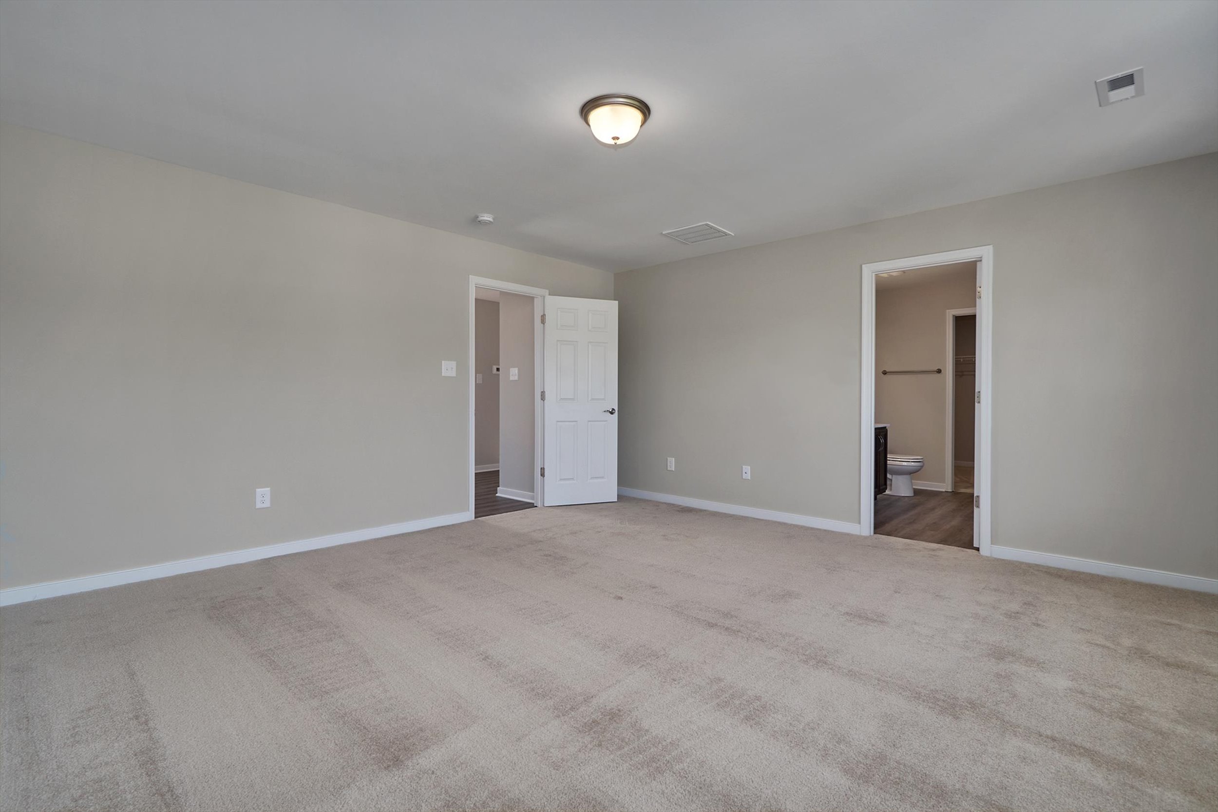 An empty room with beige carpet, light gray walls, a ceiling light fixture, and two doorways, one leading to a bathroom and the other to a hallway.