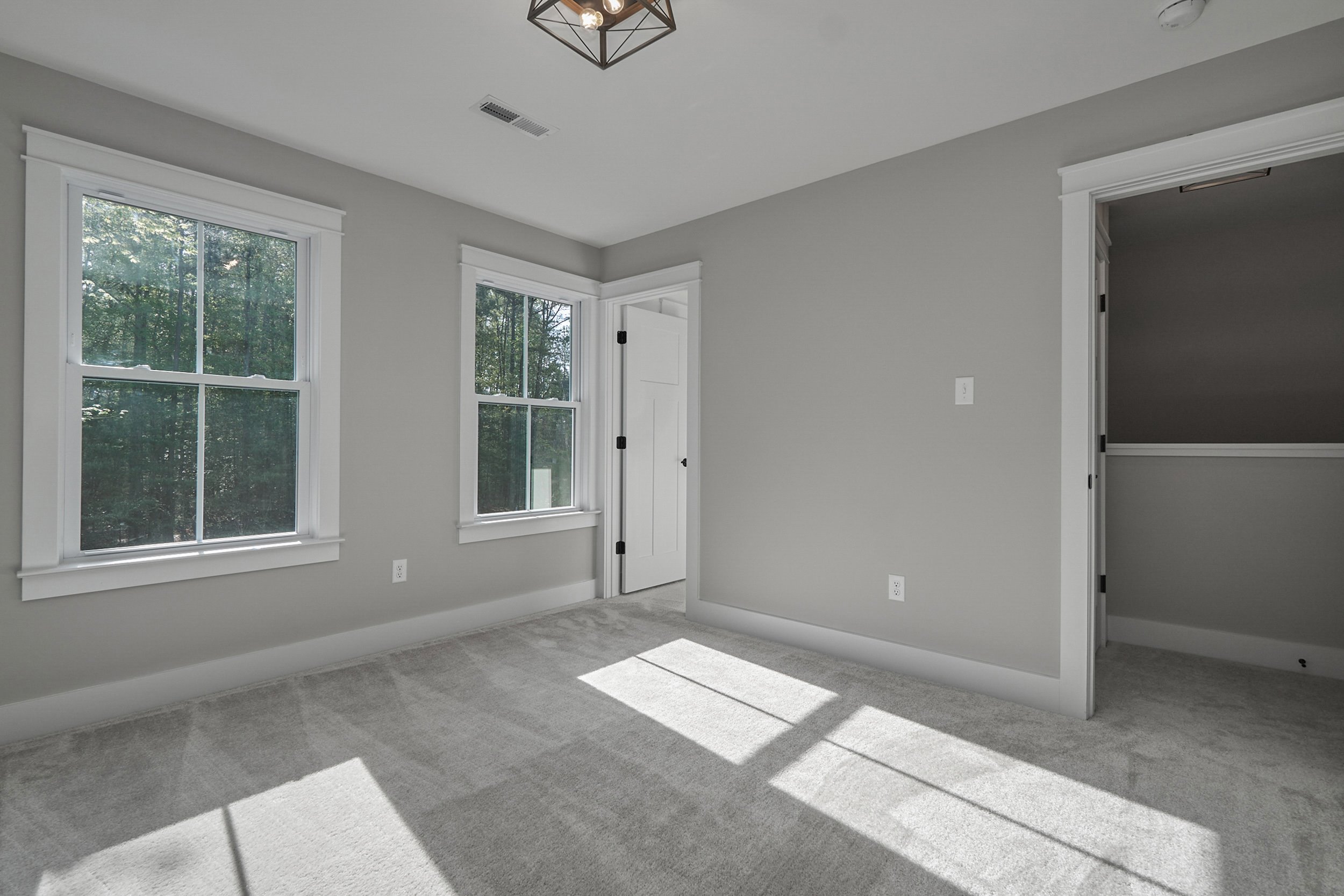A bright, empty room with grey walls, two windows, a light fixture, and a closed white door. Sunlight is streaming in through the windows, illuminating the room.