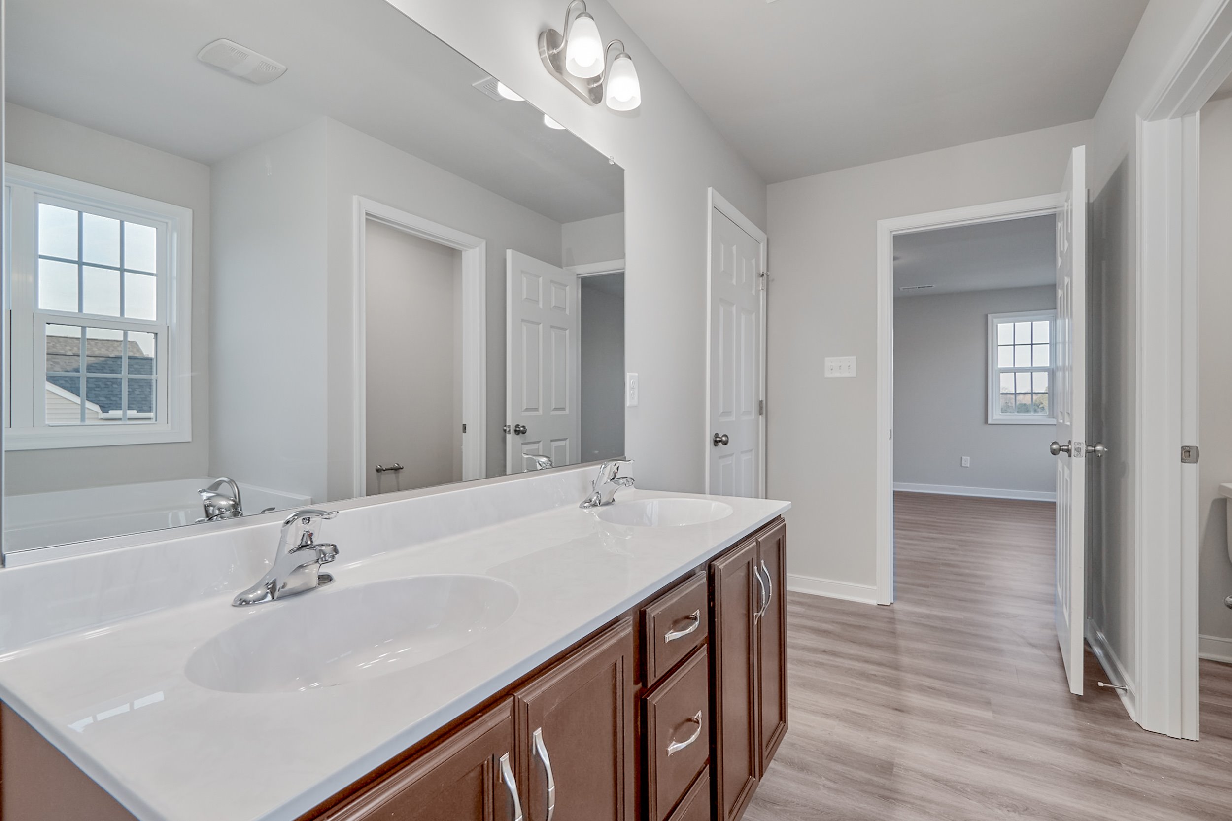 A bathroom with a double sink vanity, large mirror, and overhead lighting. The room has light-colored walls, a window, and an adjacent doorway leading to another room with wooden flooring.