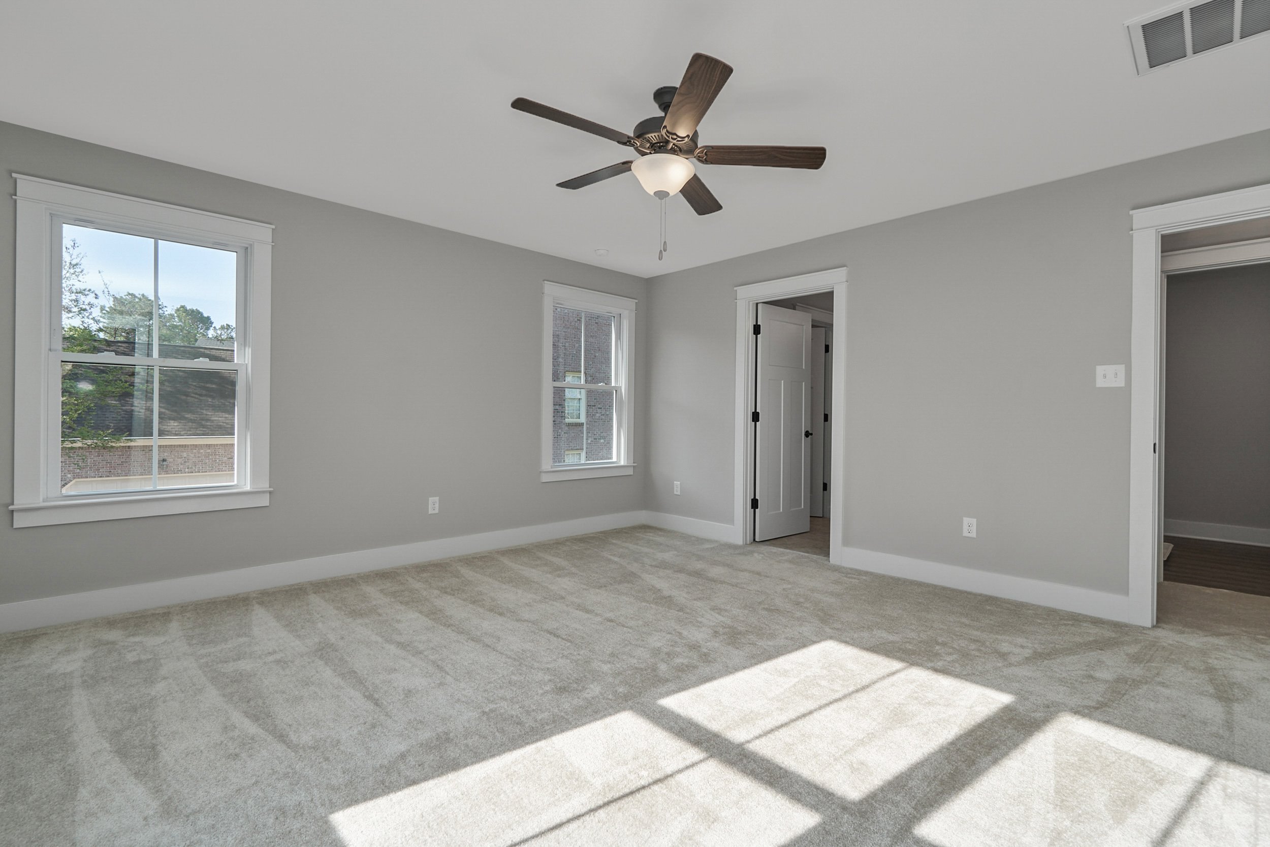 Empty, well-lit bedroom with gray walls, beige carpet, two windows, a ceiling fan, and an open door leading to an adjacent room.