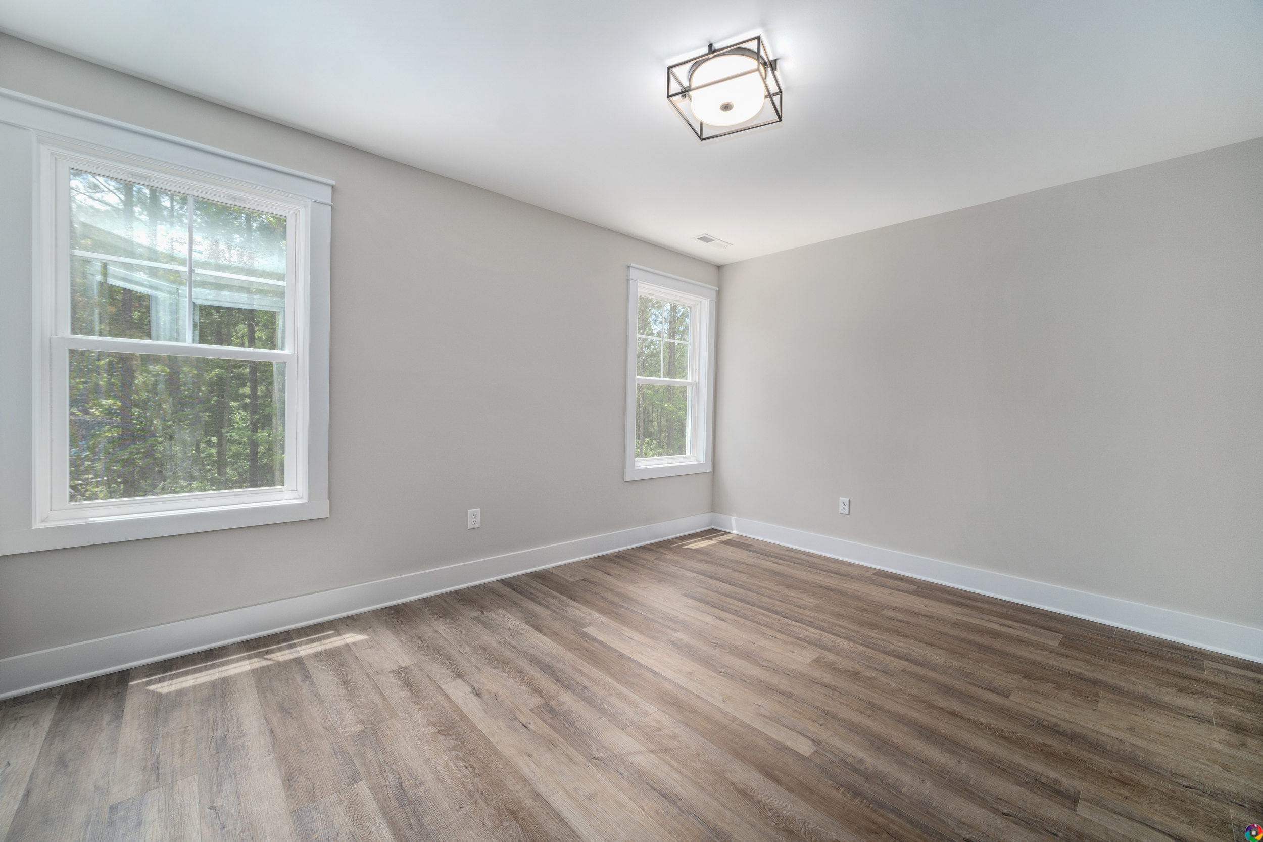 A bright, empty room with light gray walls, two large windows, wood flooring, and a modern ceiling light fixture.