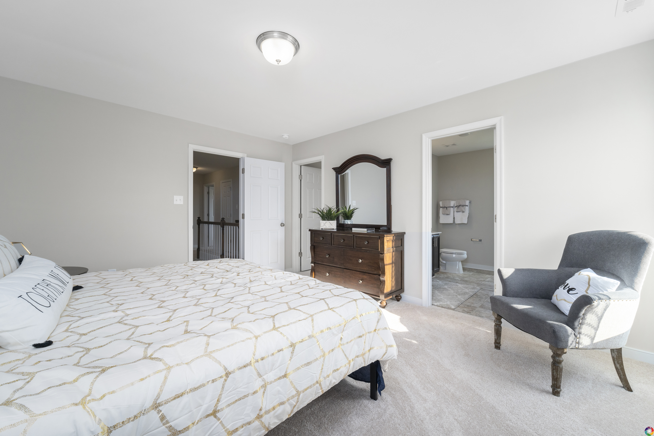 A bedroom with a bed, a wooden dresser with a mirror, a gray armchair, and an adjacent bathroom. The bed has white and gold bedding, and the walls and carpet are light-colored.