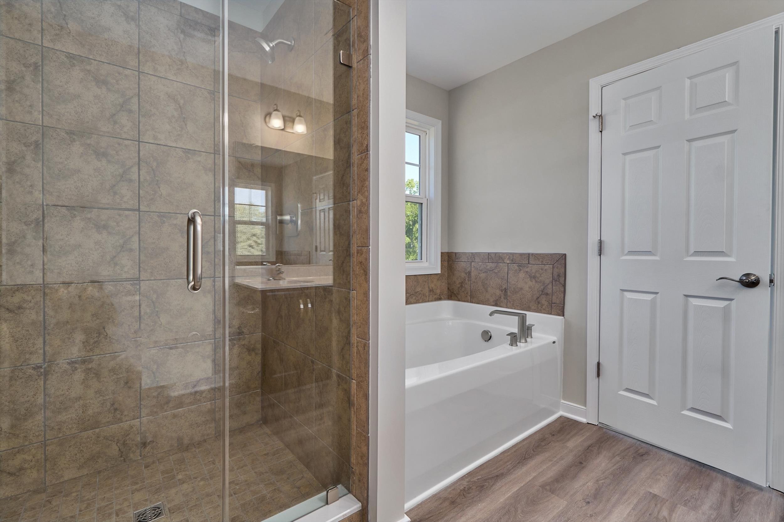 A bathroom featuring a glass-enclosed shower with beige tiles, a white bathtub with silver fixtures, a window, and a white door with a silver handle.