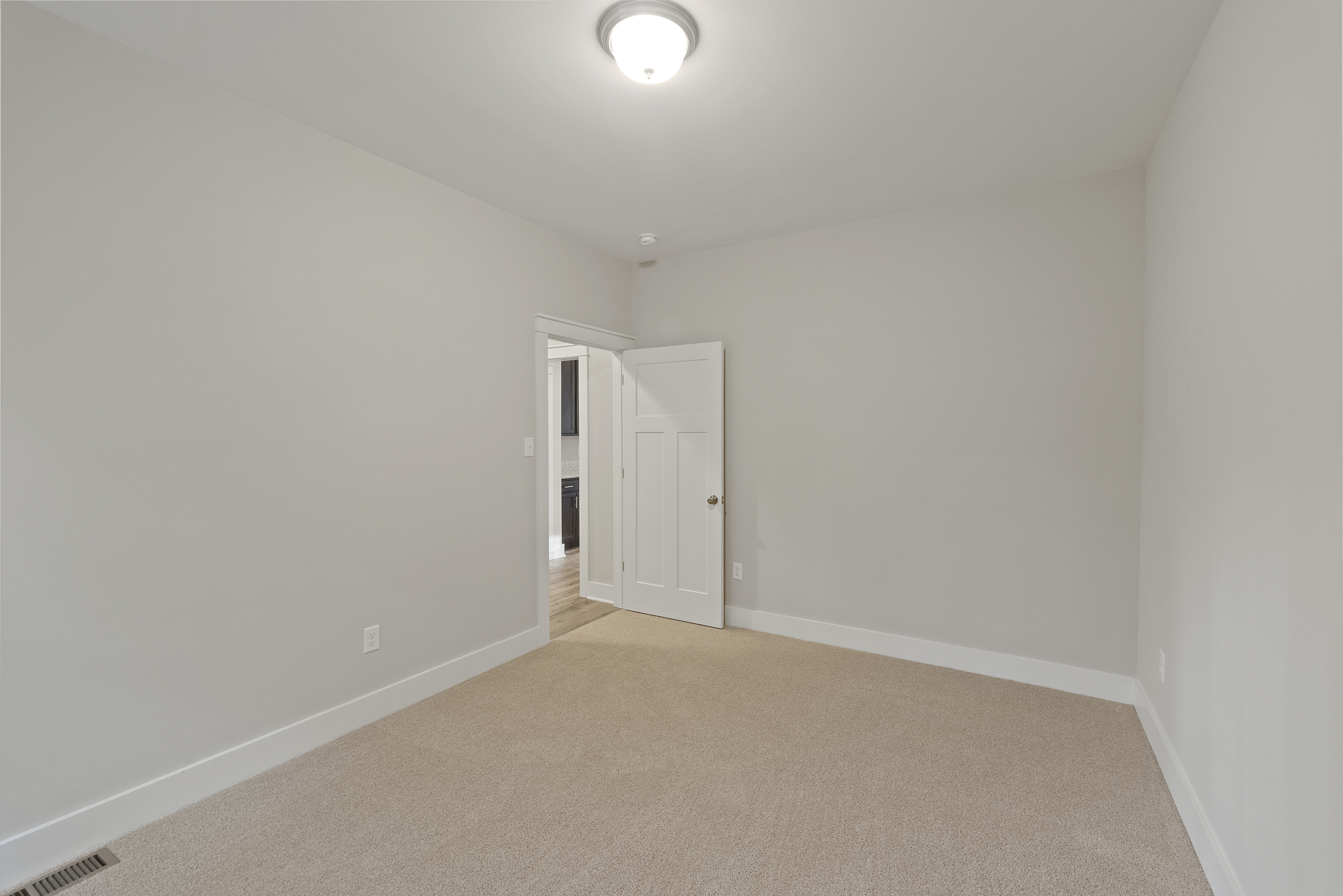 Empty room with beige carpet, light gray walls, a white ceiling light fixture, and a partially open white door leading to another room.