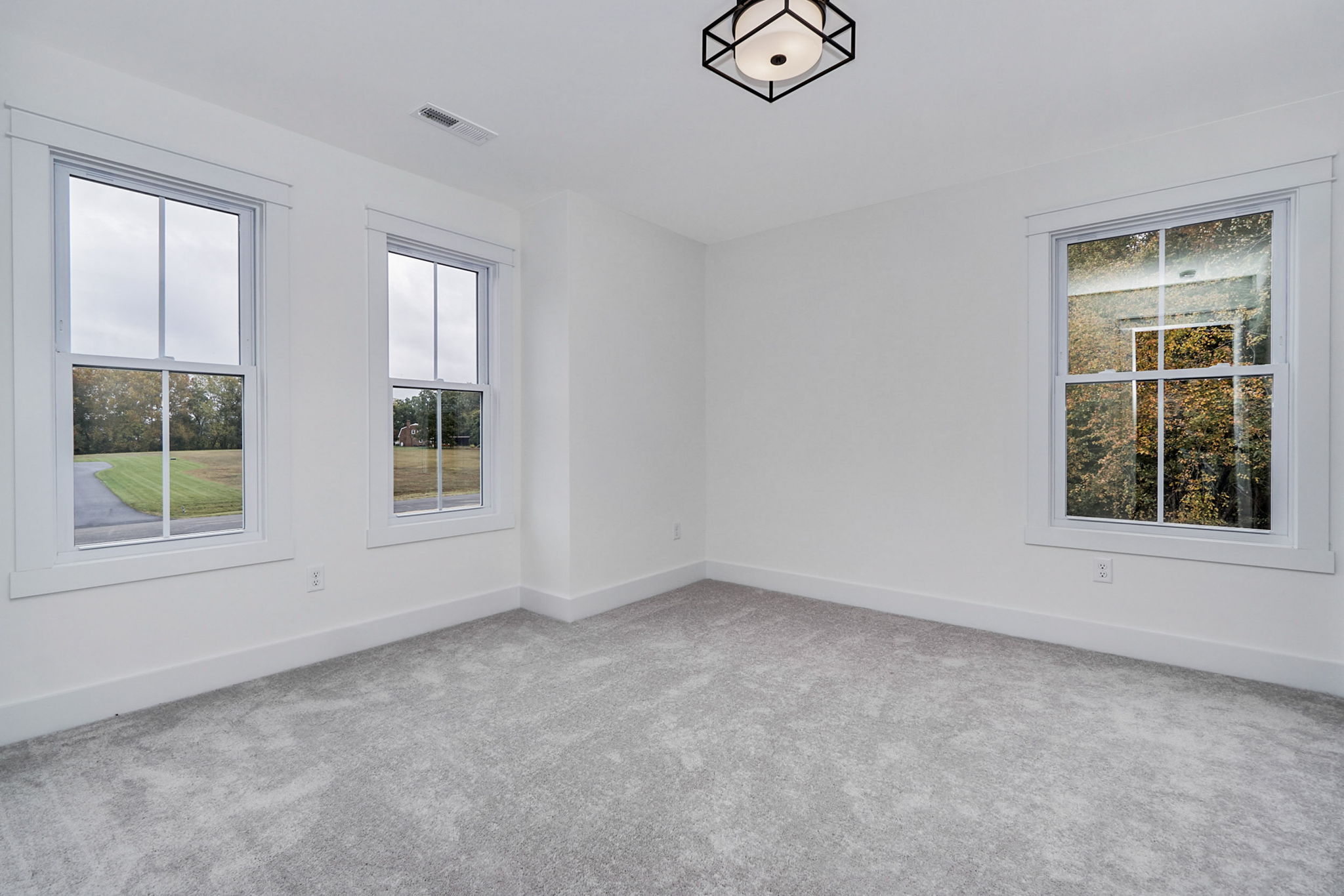 Empty room with white walls and carpeted floor, featuring two large windows on the left wall and one on the right, allowing natural light to enter. A modern light fixture hangs from the ceiling.