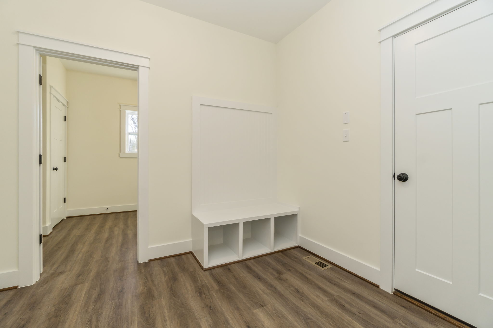 An empty room with beige walls, a white bench with storage cubbies, and two closed doors leading to other rooms.