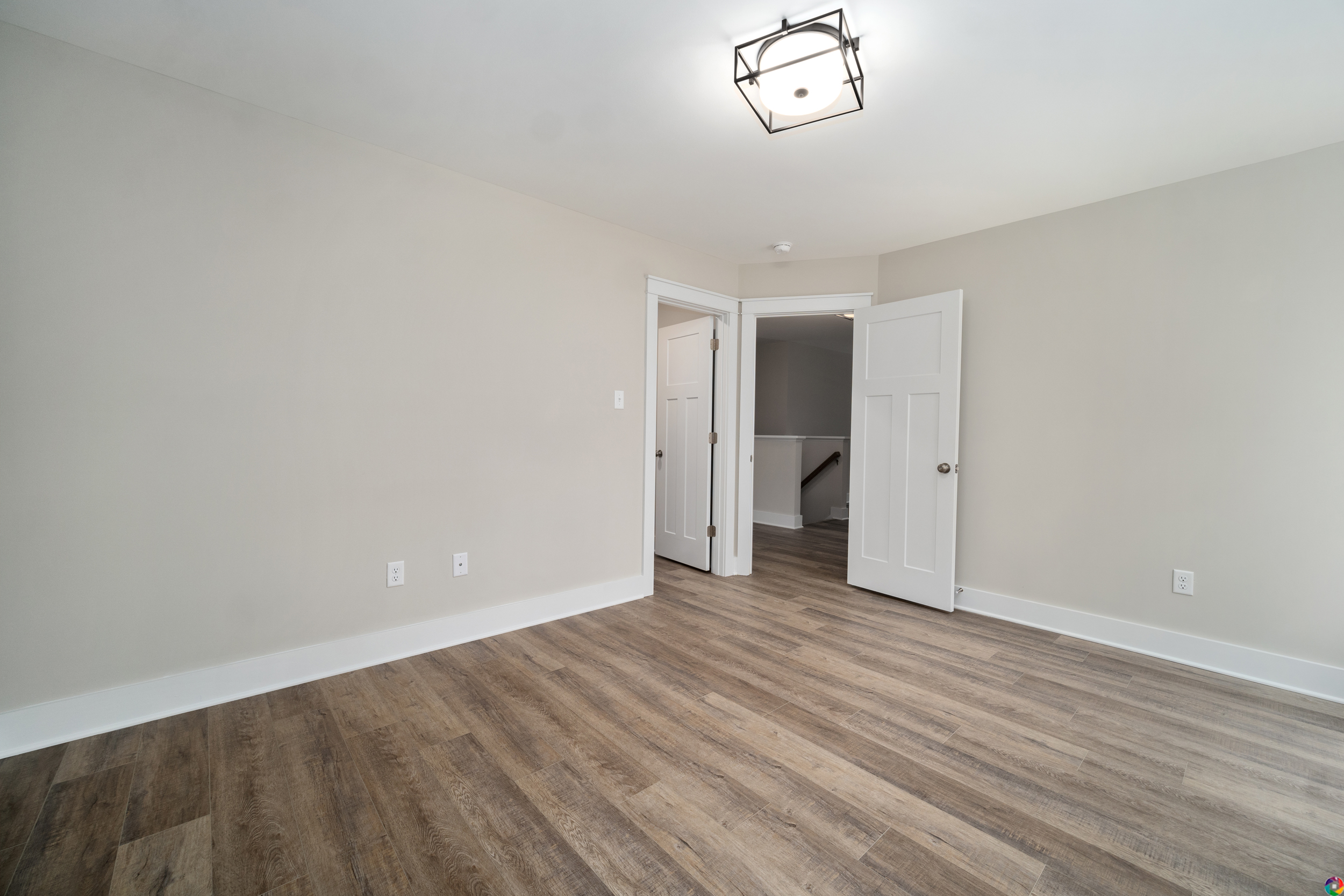 An empty, light-filled room with beige walls, wood flooring, a modern ceiling light fixture, two open doors, and white trim doors and baseboards.
