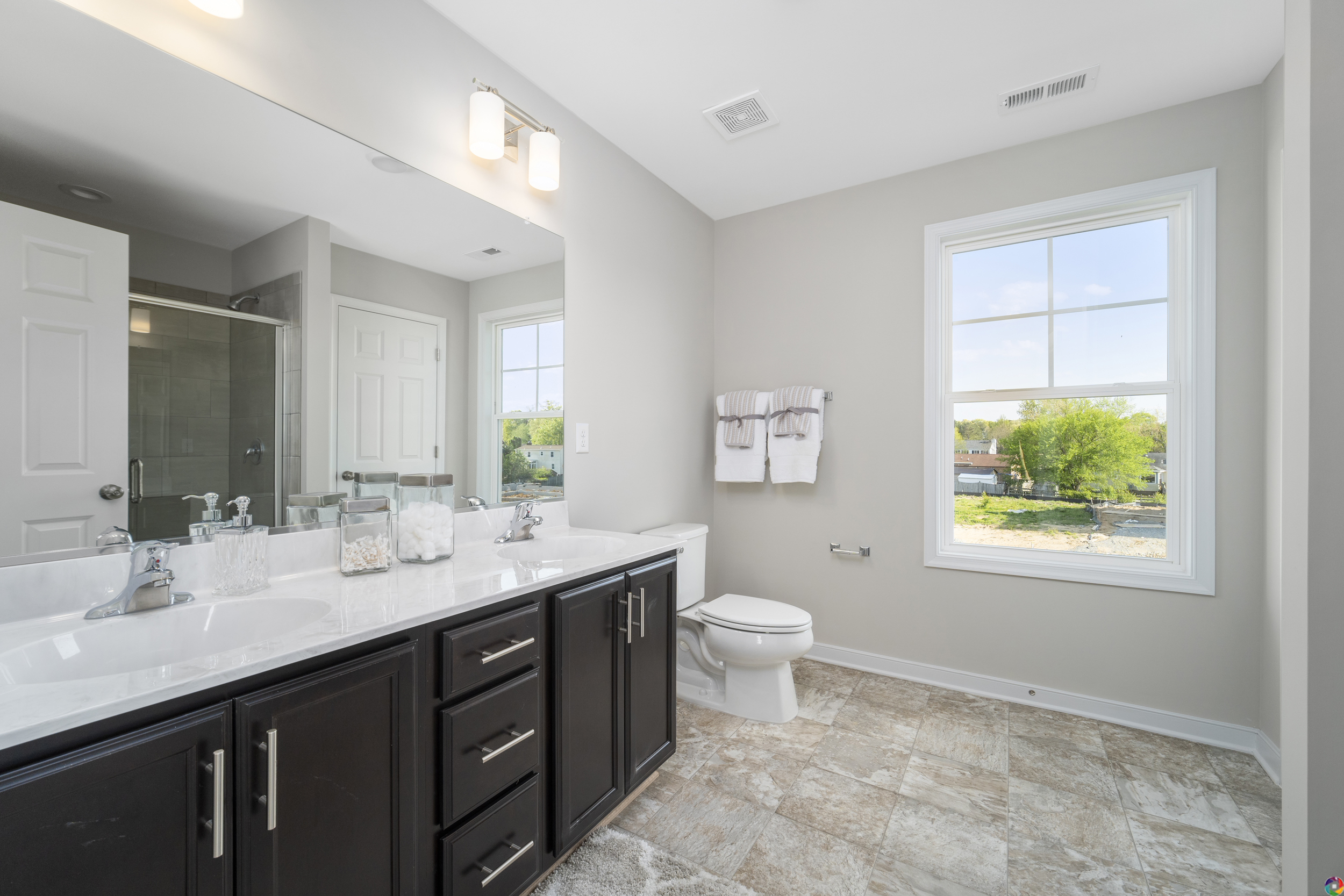 Modern bathroom with double sink vanity and large mirror on the left, toilet in the center, glass shower in the back, and a window providing natural light. Towel rack with neatly folded towels on the wall.
