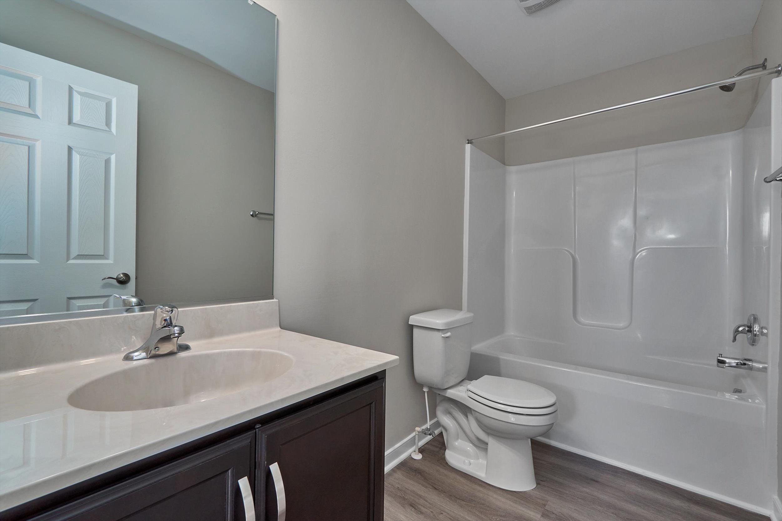 A modern bathroom with a large mirror, single sink vanity, toilet, and a shower-tub combination. The room has light gray walls and a wood-style floor.