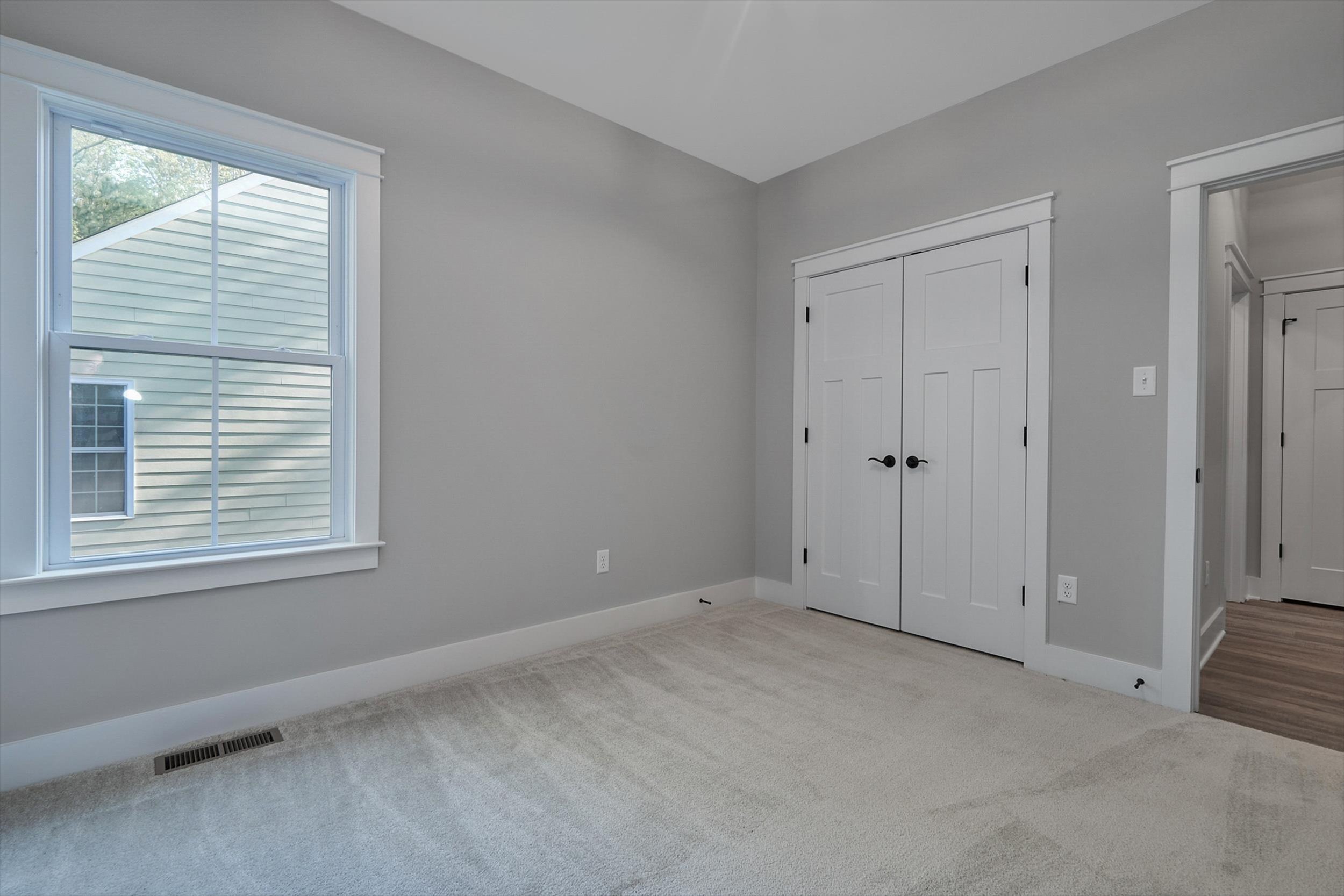 A simple, empty room with gray walls, beige carpet, a window, and double-door closet. There are open doors to the hallway visible on the right.