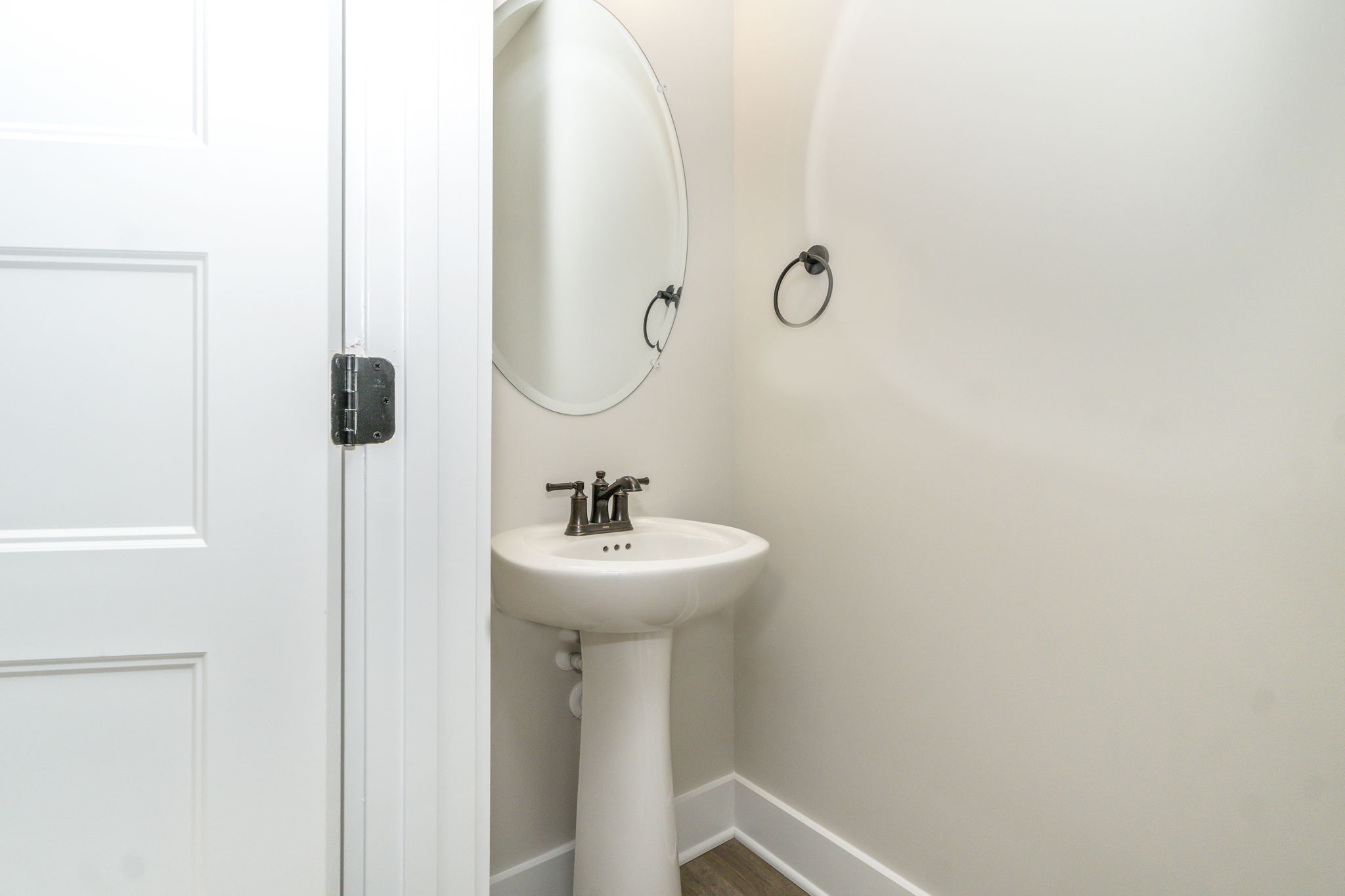 A small bathroom with a white pedestal sink, round mirror, and an open door on the left.