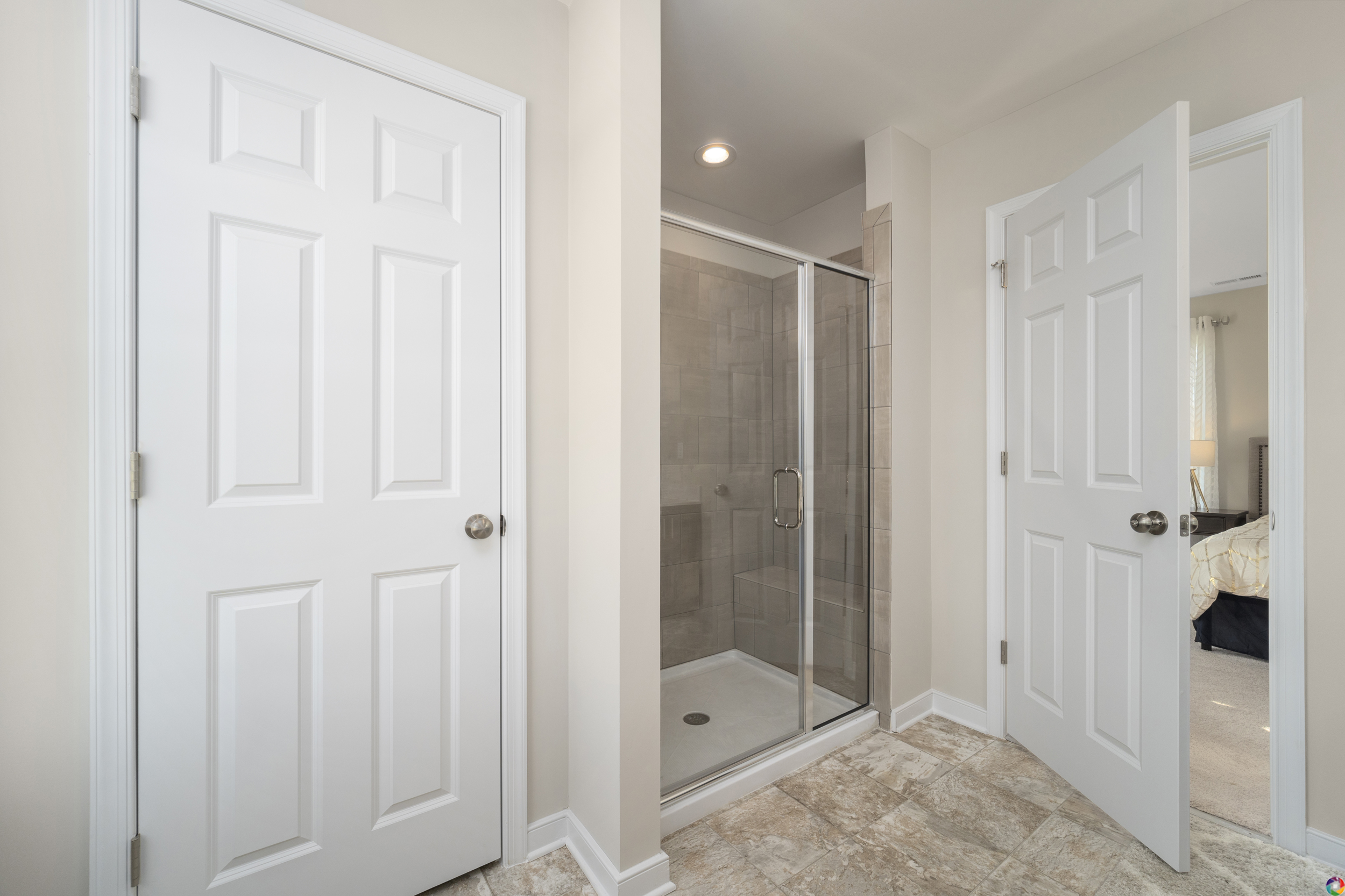 A bathroom featuring a tiled shower with a glass door. There are two white doors on either side of the shower area, and the floor is tiled.