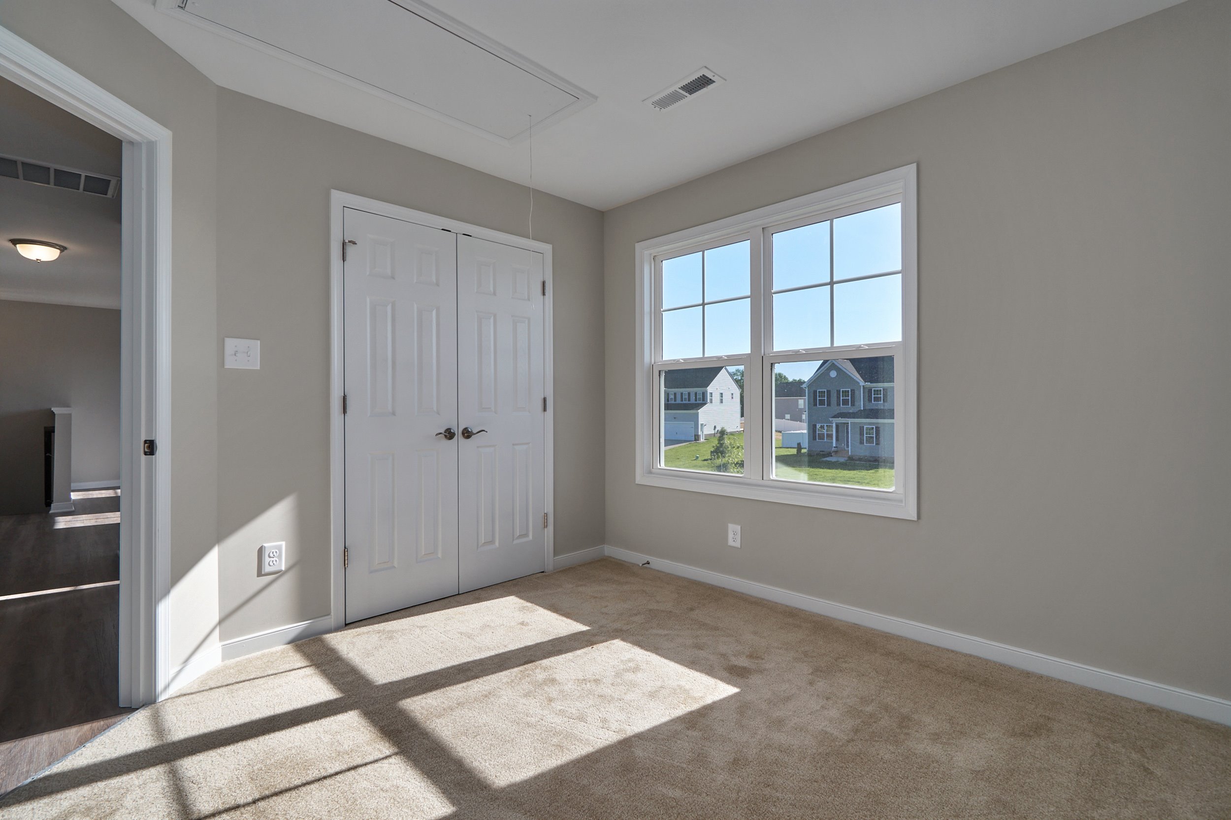 A small, empty bedroom with beige walls and carpet, a window with a view of neighboring houses, and a closed double-door closet.