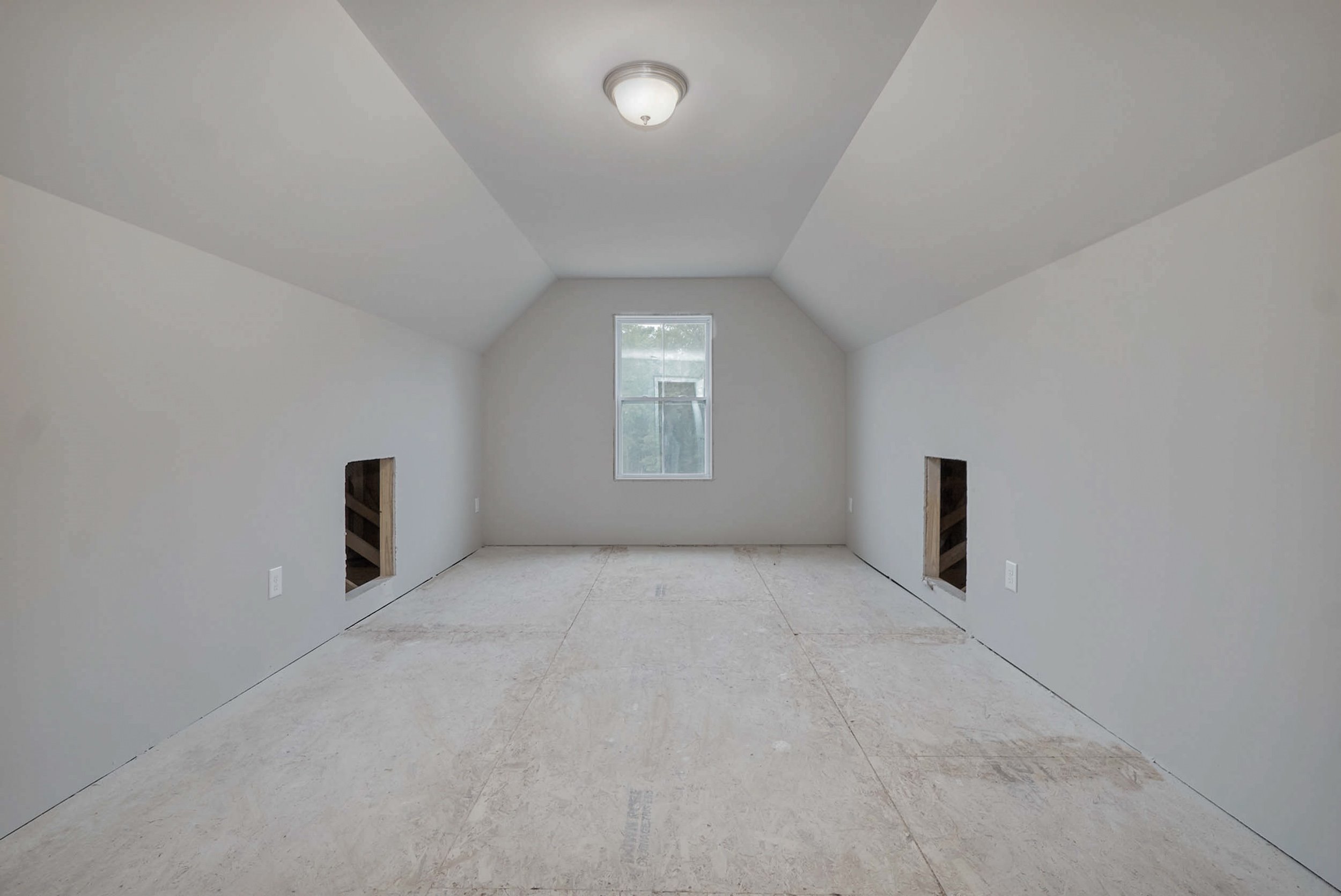 An unfinished attic room with slanted walls, a ceiling light, a central window, and two small alcoves on either side. The floor is bare, and the walls are painted white.