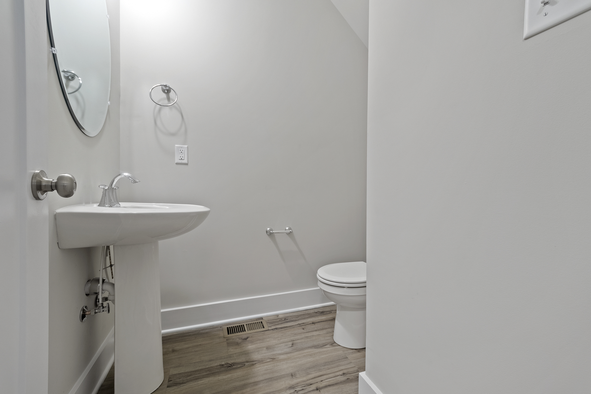 A small bathroom with a pedestal sink, round mirror, towel ring, toilet, and white walls. The floor has light wood-like flooring, and there is a wall vent near the baseboard.