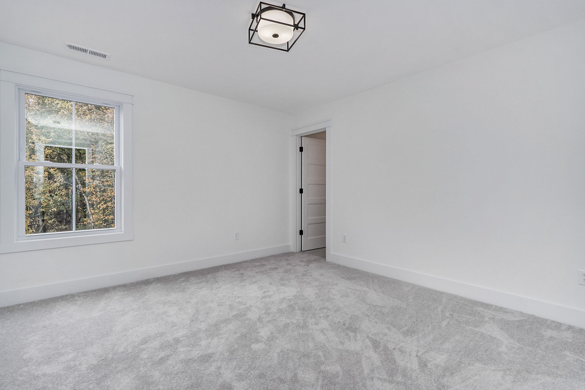 Empty room with light gray carpet, white walls, a single window on the left, and a door adjacent to the window. Ceiling has a geometric black light fixture.
