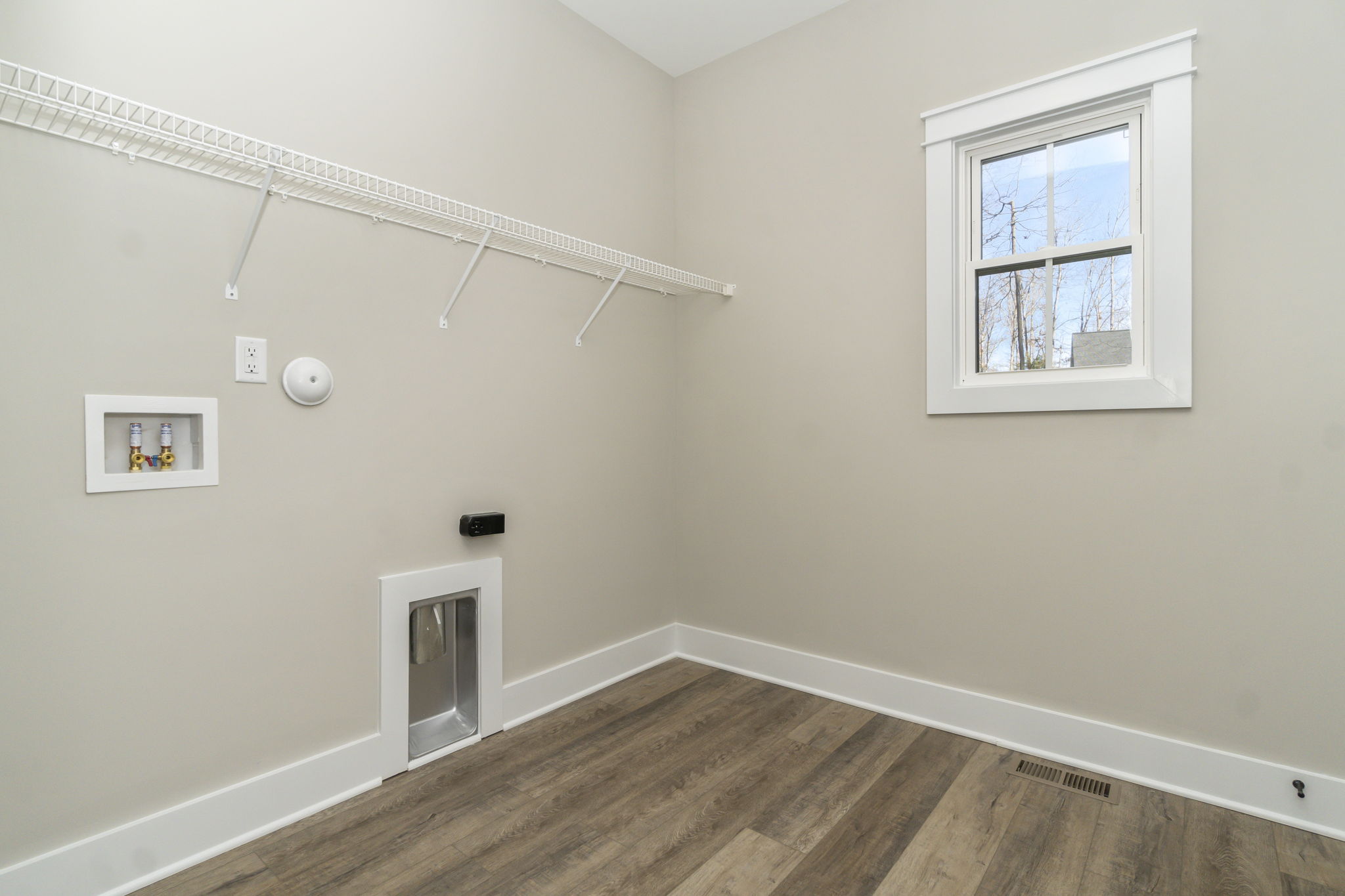 A small, empty room with a window, gray walls, a white door with a built-in pet entrance, and a wall-mounted wire shelf.