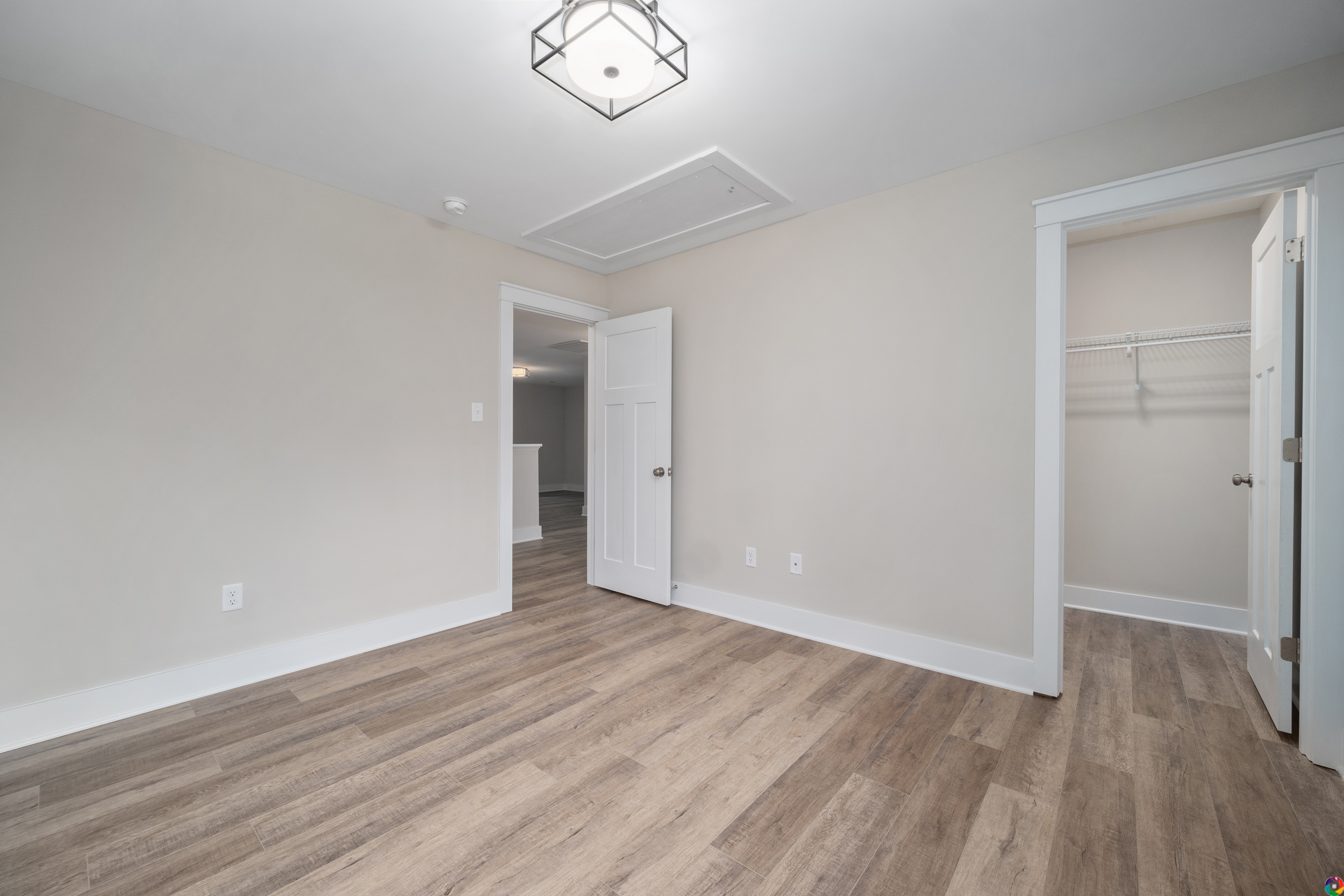 An empty room with light gray walls, wood flooring, a white door, a closet with shelves, and a modern ceiling light fixture.