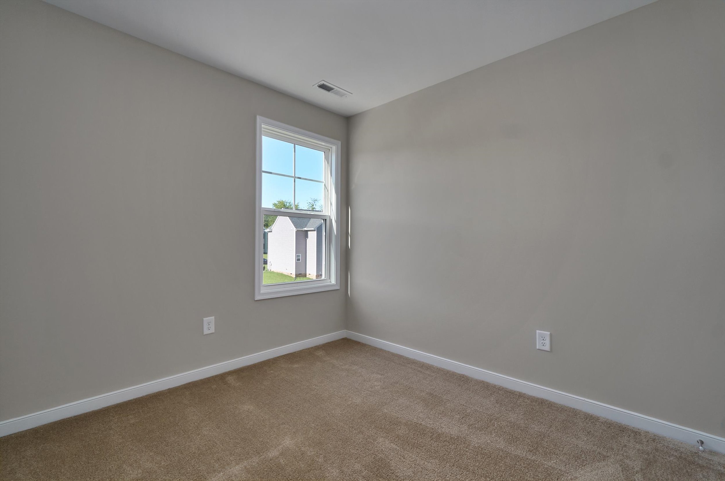 Empty room with beige carpet, light gray walls, and a single window showing an outside view of buildings. The room has an electrical outlet on one wall.