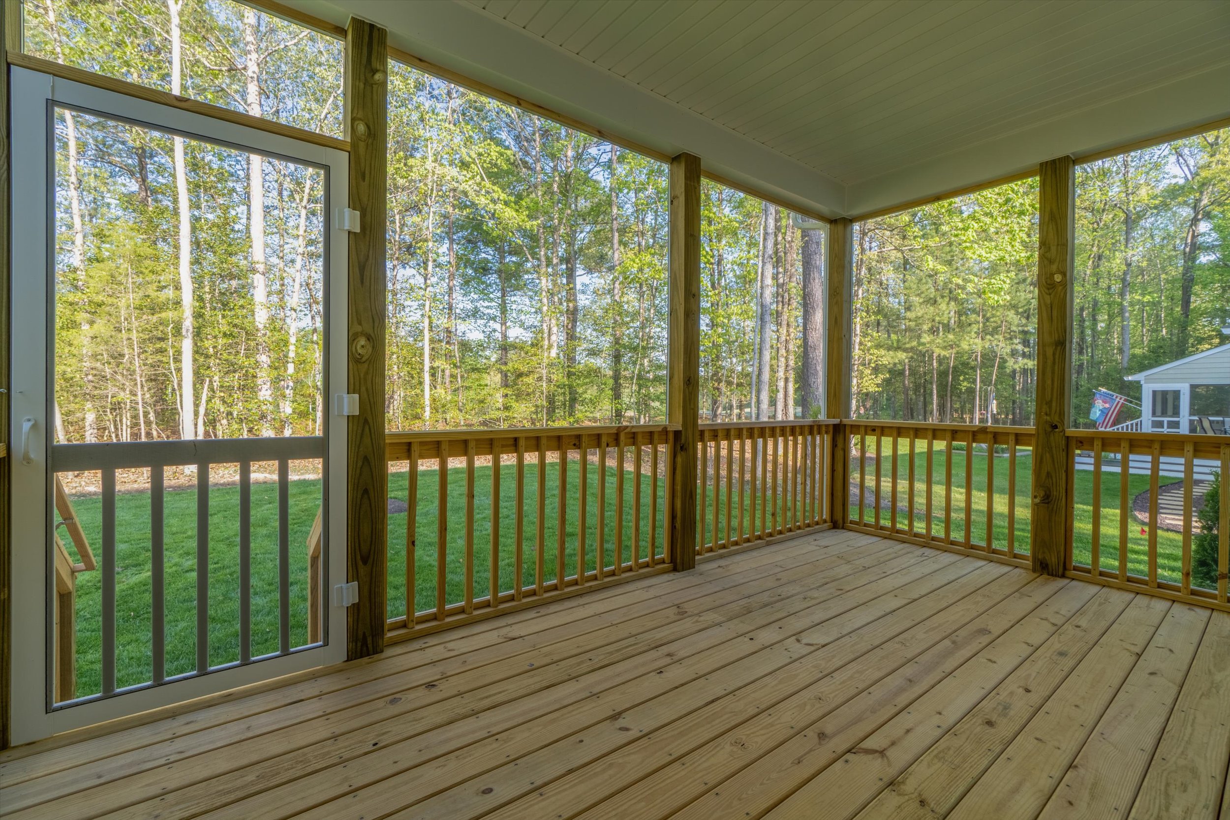 A wooden deck with a screened door overlooks a grassy lawn and wooded area in the background.