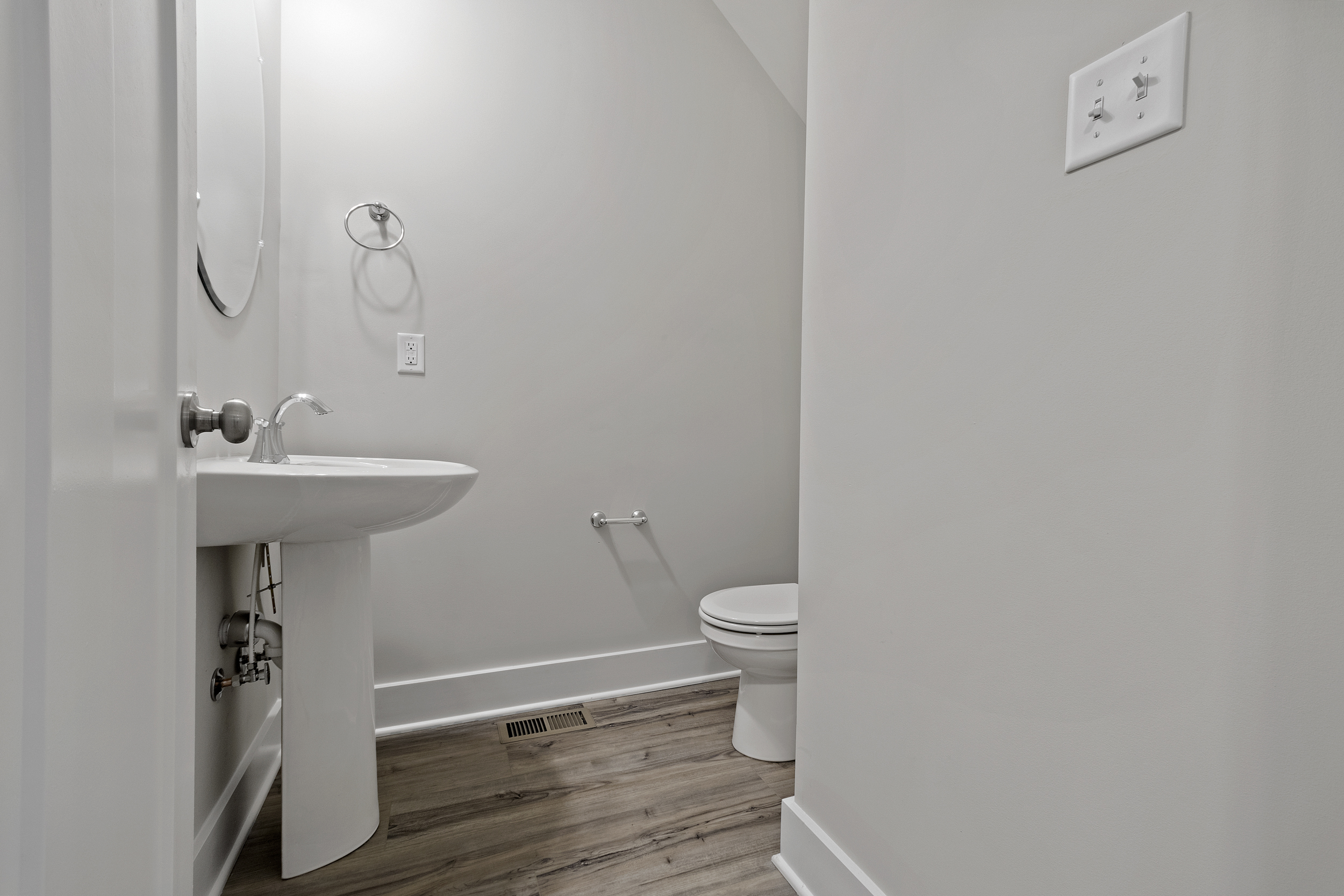A small bathroom with a pedestal sink, wall-mounted faucet, circular mirror, toilet, and light grey walls with wood-look floor tiles.