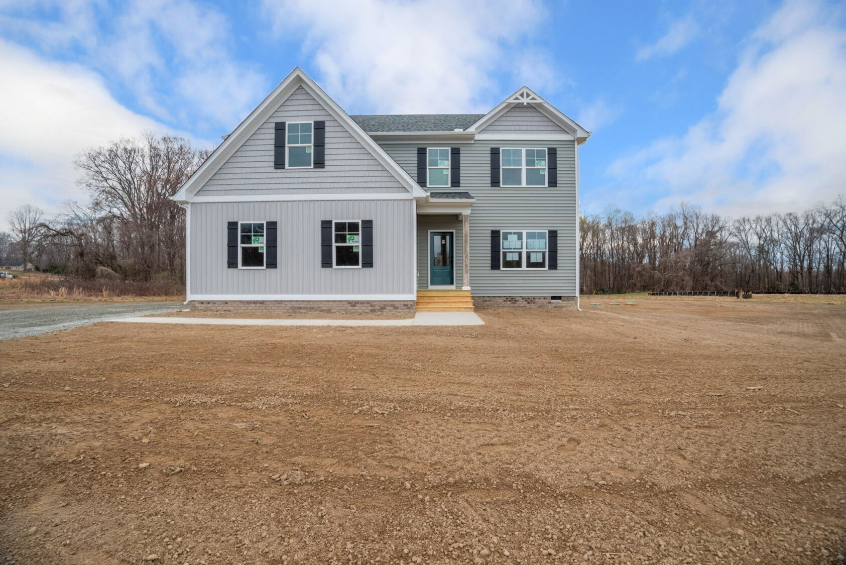 A newly constructed two-story house with gray siding, black shutters, a dark green front door, and multiple windows, surrounded by an unfinished yard with dirt ground, under a partly cloudy sky.