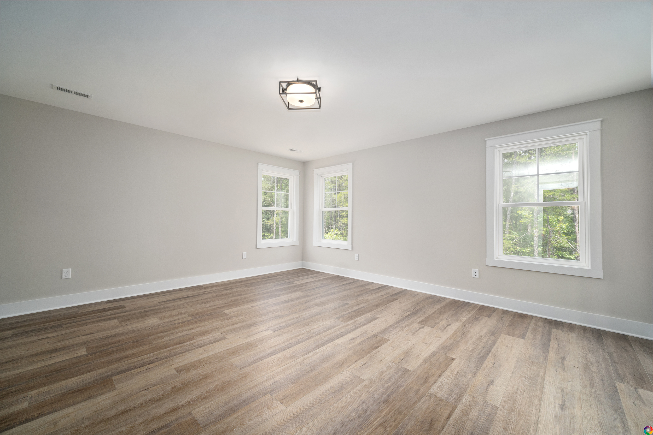 An empty, light-filled room with wooden flooring, beige walls, a square ceiling light, and three windows displaying greenery outside.