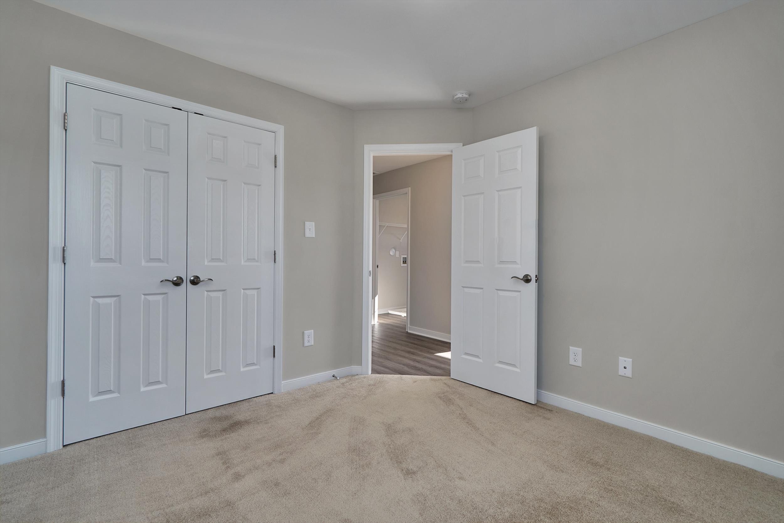 A beige carpeted room with two white closet doors on the left and a white open door on the right leading to a hallway with wooden floors. Walls are painted light gray.