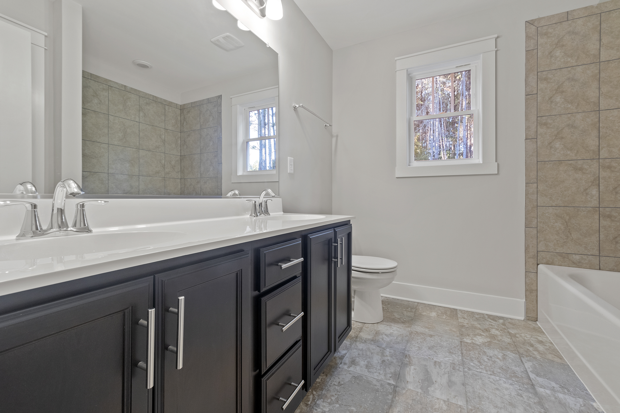 Modern bathroom featuring a dark wood double vanity with a large mirror, dual sinks, a toilet, a bathtub, tiled floors and walls, and a window with a view of trees.