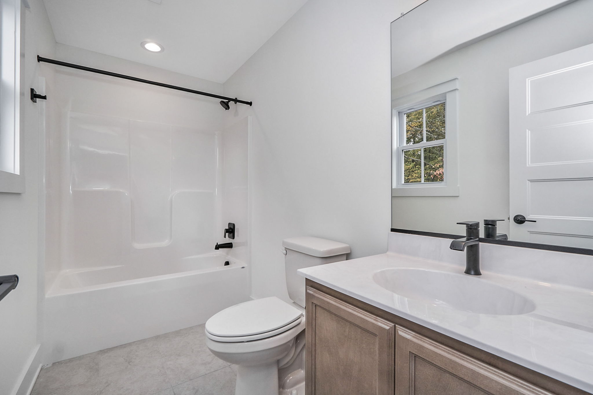 Clean, modern bathroom with a white bathtub, toilet, and sink. The room features a large mirror, light gray flooring, and a small window near the tub.