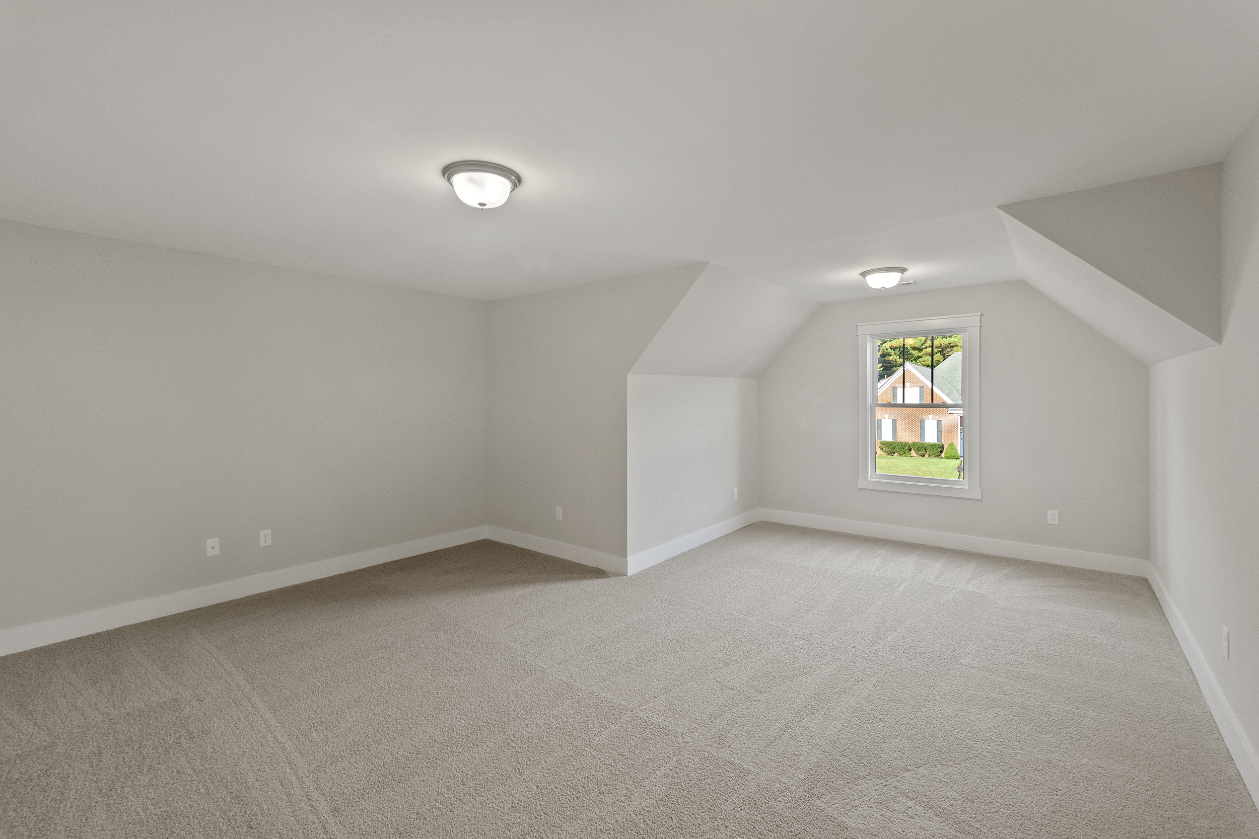 A spacious, empty room with light gray walls, beige carpet, two ceiling lights, and a single window showing a view of a house.