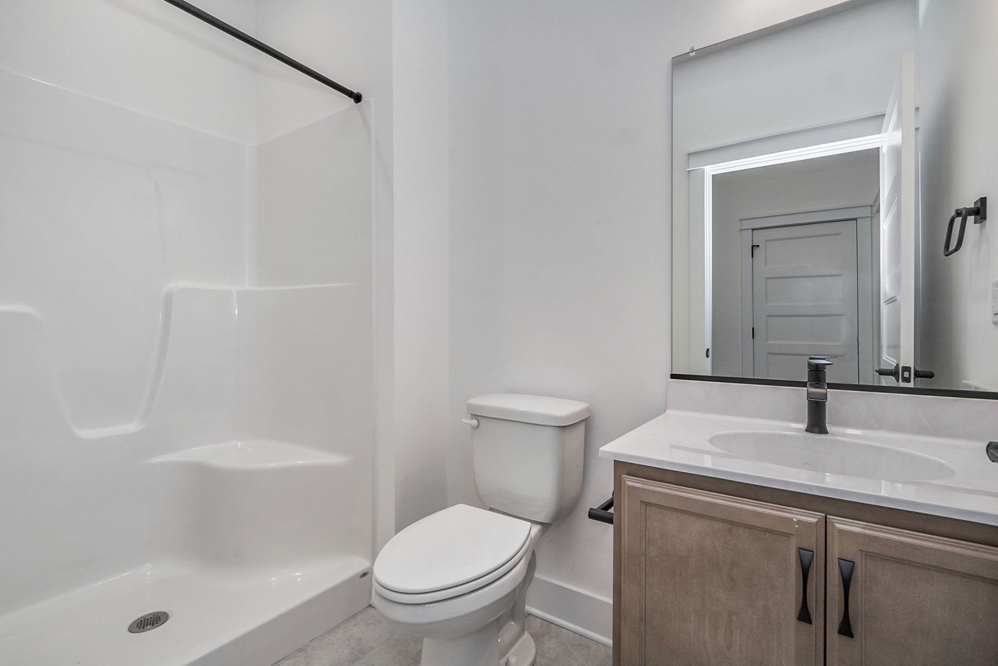 A clean, modern bathroom features a white shower enclosure, a toilet, and a vanity with a sink underneath a large mirror. Minimalistic design with a neutral color scheme.