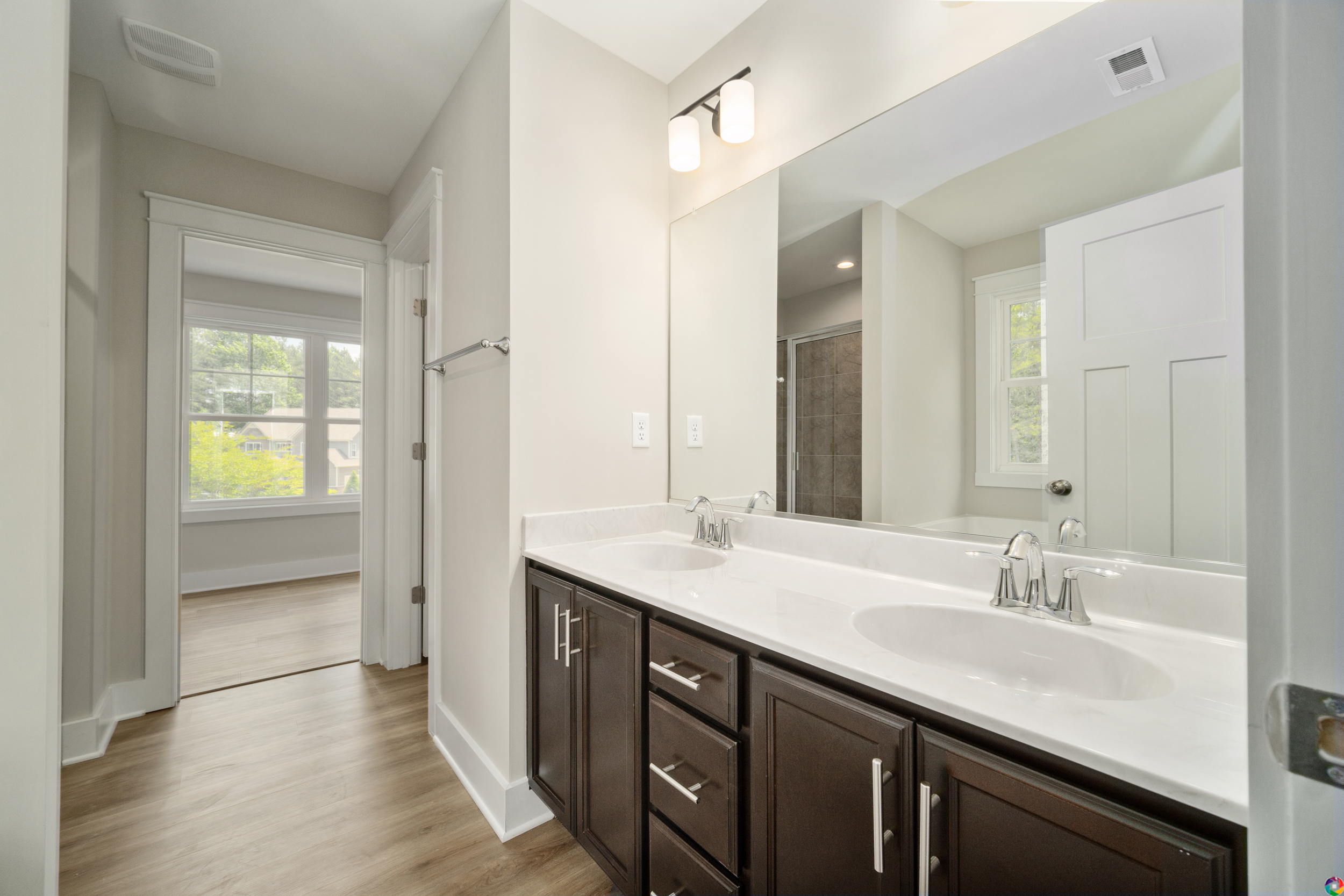 A modern bathroom featuring a double sink vanity with dark cabinets, a large mirror, bright lighting, and an adjacent room visible in the background with a large window.